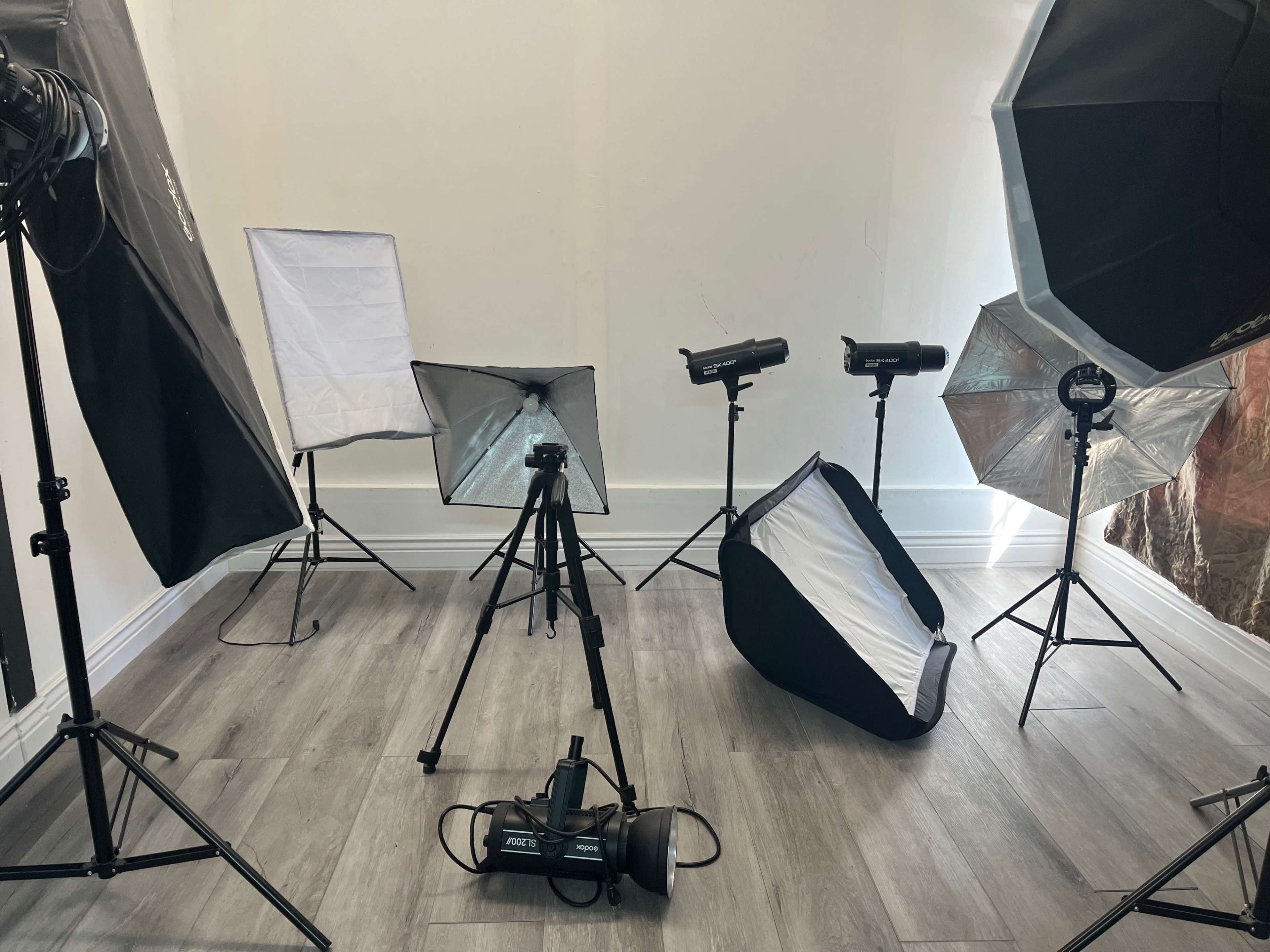 The image shows a photography studio setup with various lighting equipment, softboxes, and a tripod arranged around a light-colored backdrop.