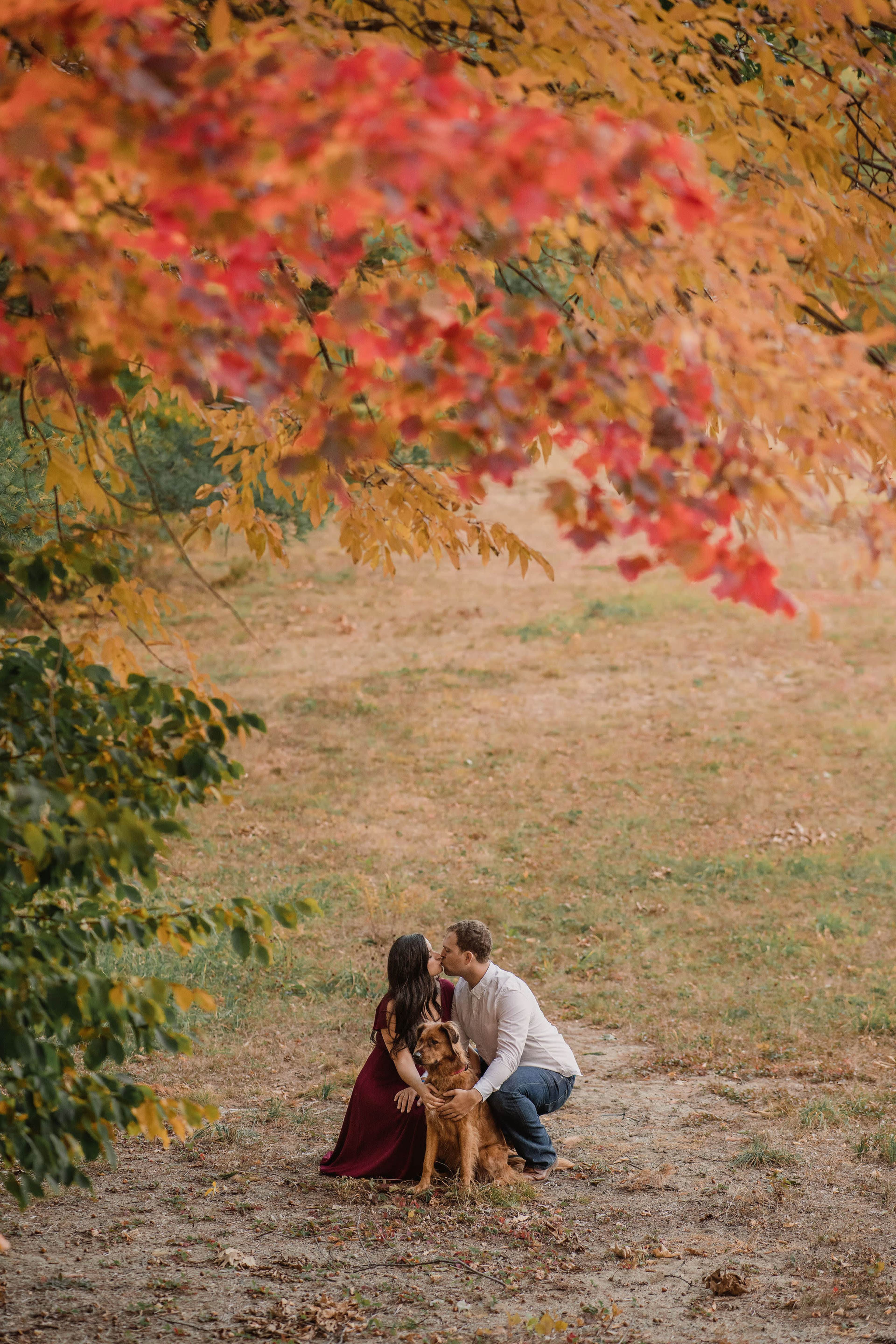 A couple kneels together in a grassy area surrounded by fall foliage, while a dog sits nearby.