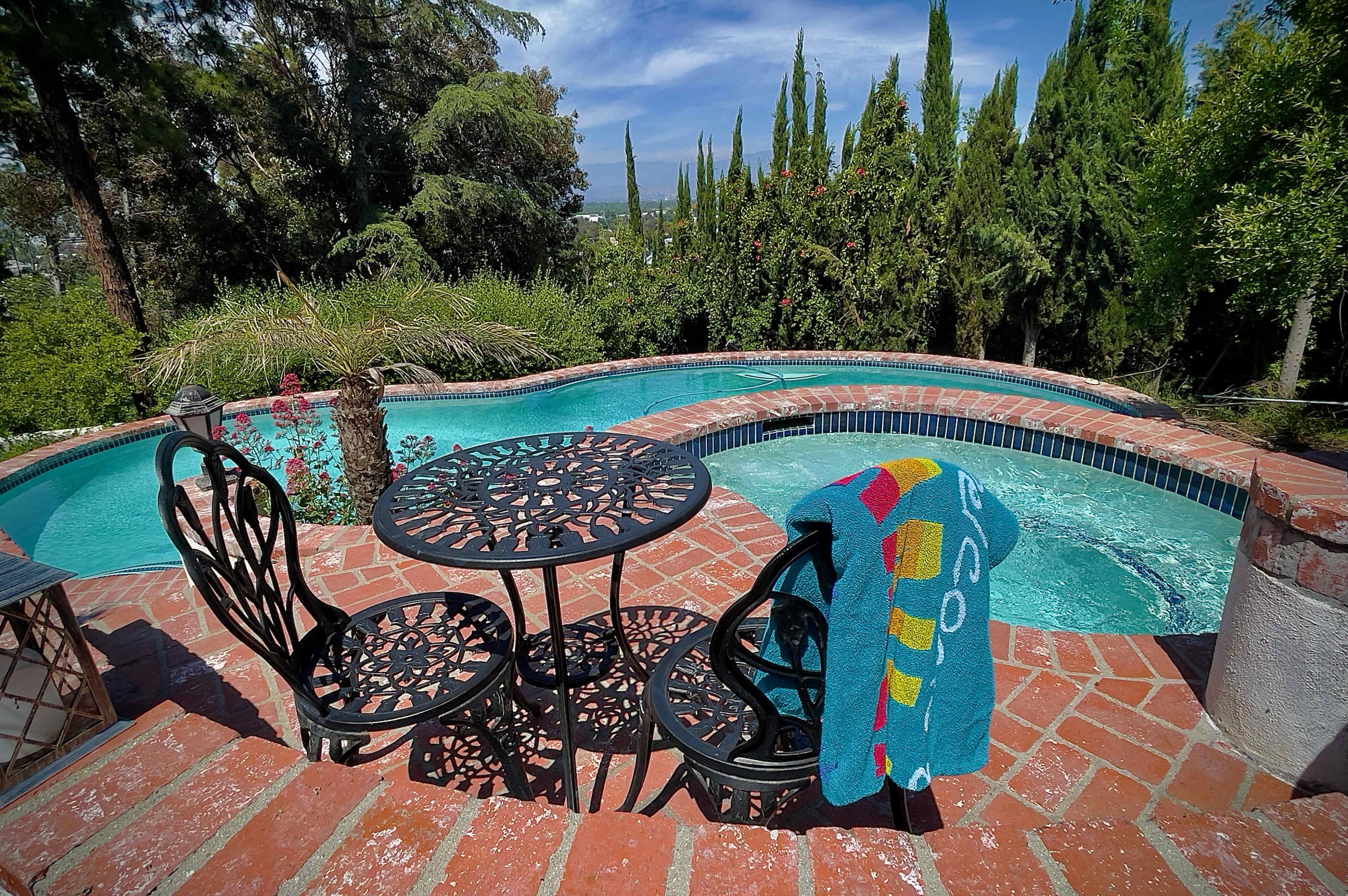 A circular patio table and two chairs are positioned next to a swimming pool surrounded by lush greenery.
