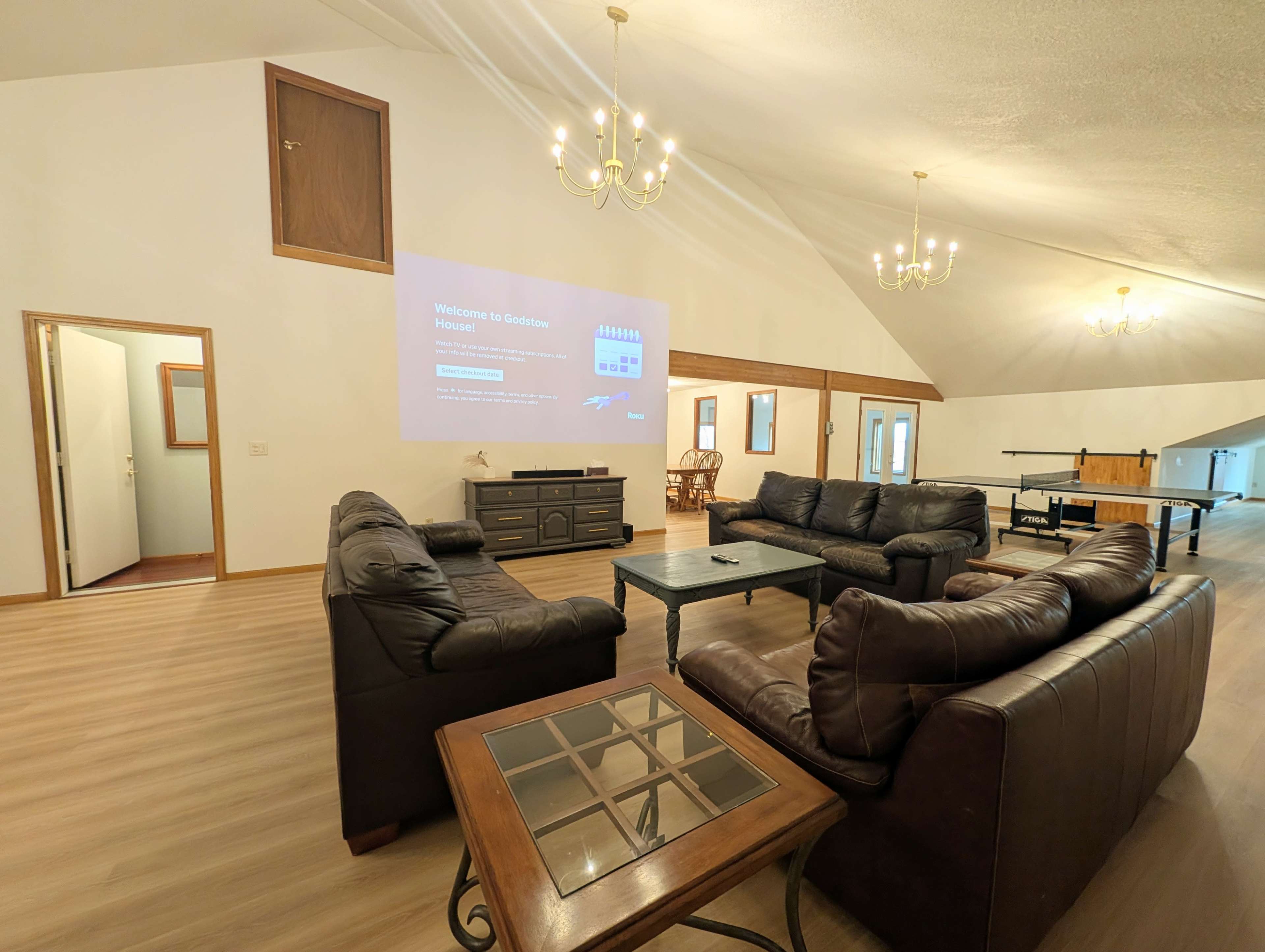 The image shows a spacious room with multiple seating areas, featuring brown leather sofas, a coffee table, and a large projection screen on one wall.
