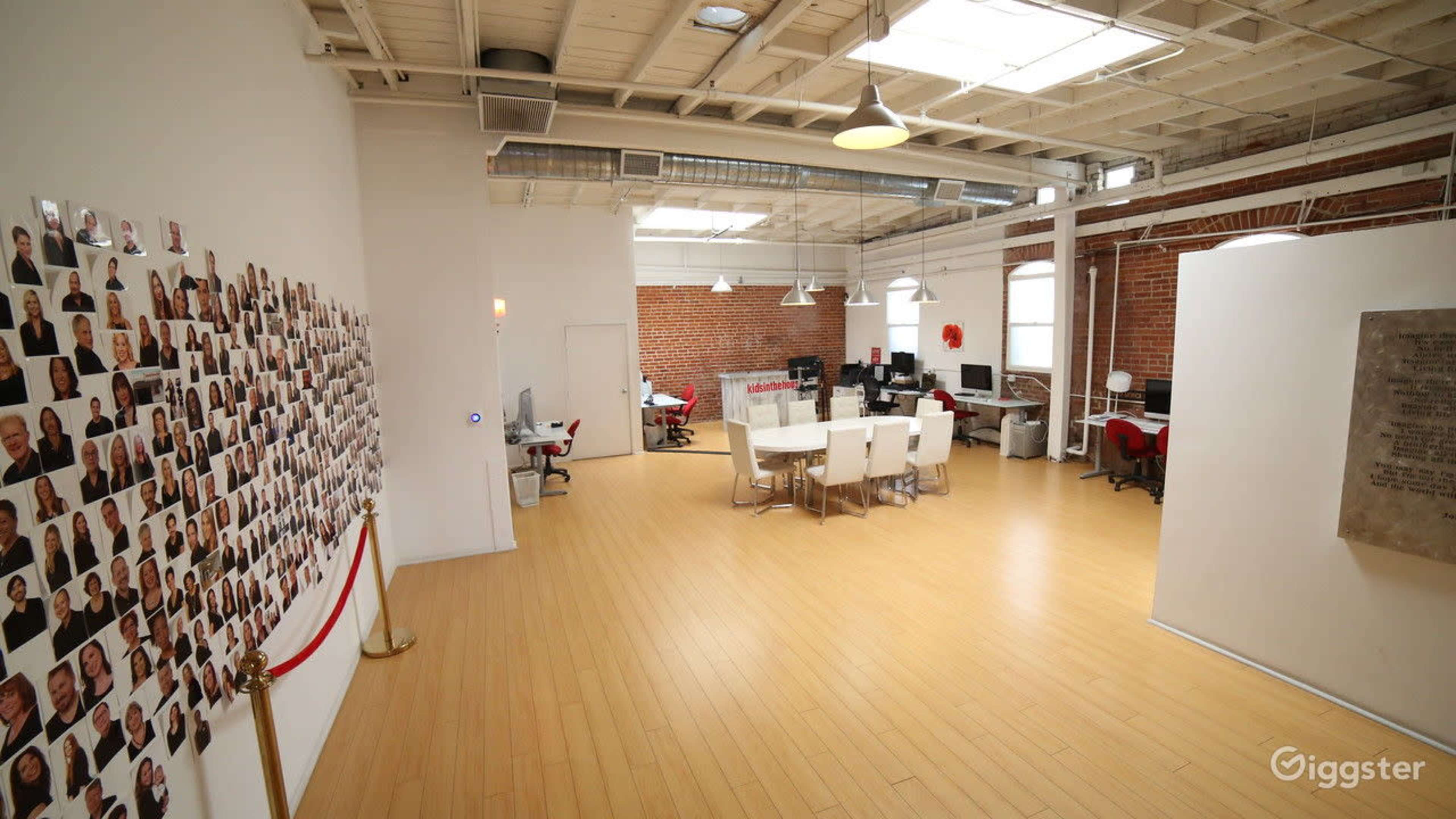 A large modern office space with a polished wooden floor, featuring a long table surrounded by white chairs, a wall covered with numerous photographs, and exposed brick walls.