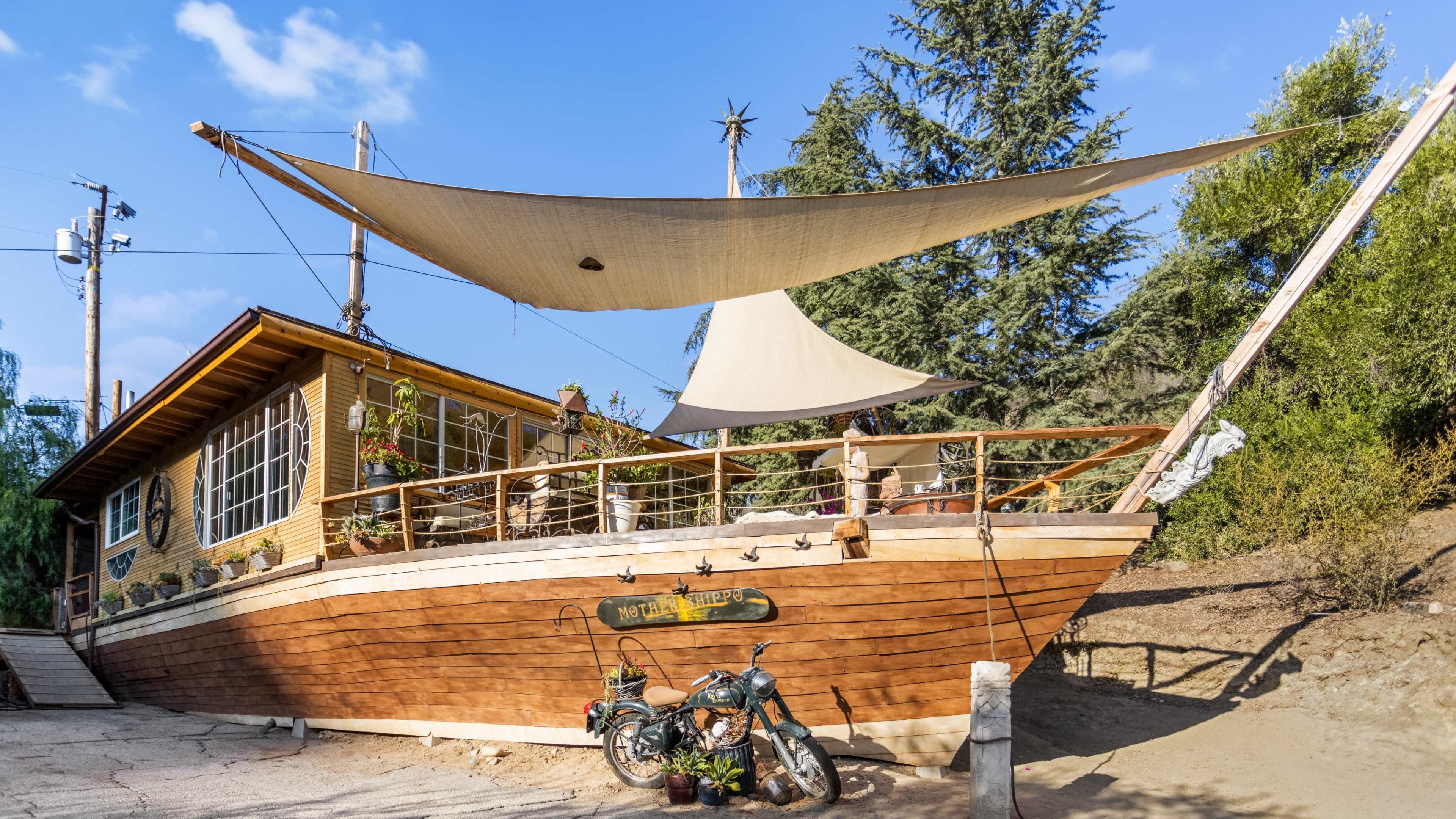 A wooden house designed to resemble a ship sits on a slope, with a large sail covering the deck and a motorcycle parked nearby.