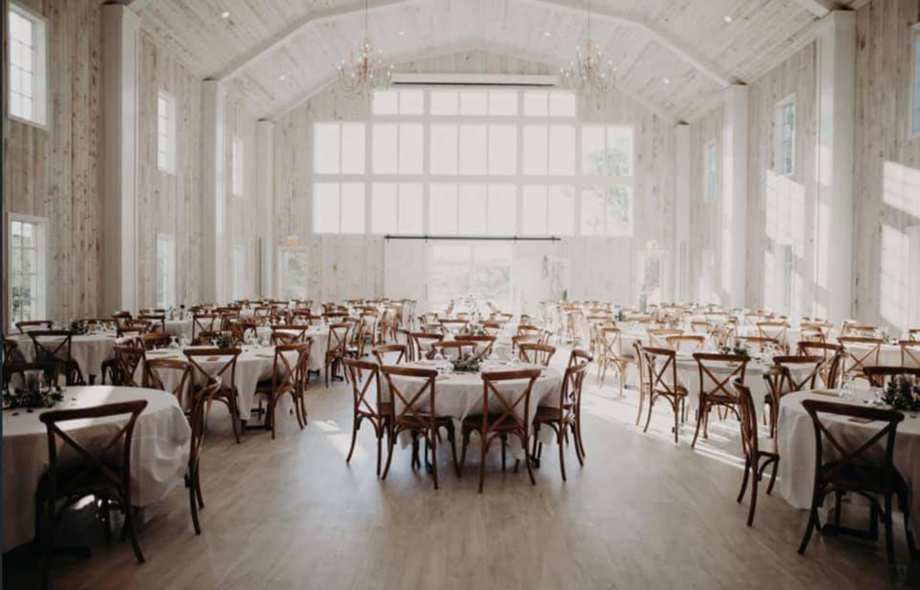 The image depicts a spacious event hall with wooden walls, multiple round tables set with white tablecloths, and large windows allowing natural light to fill the room.