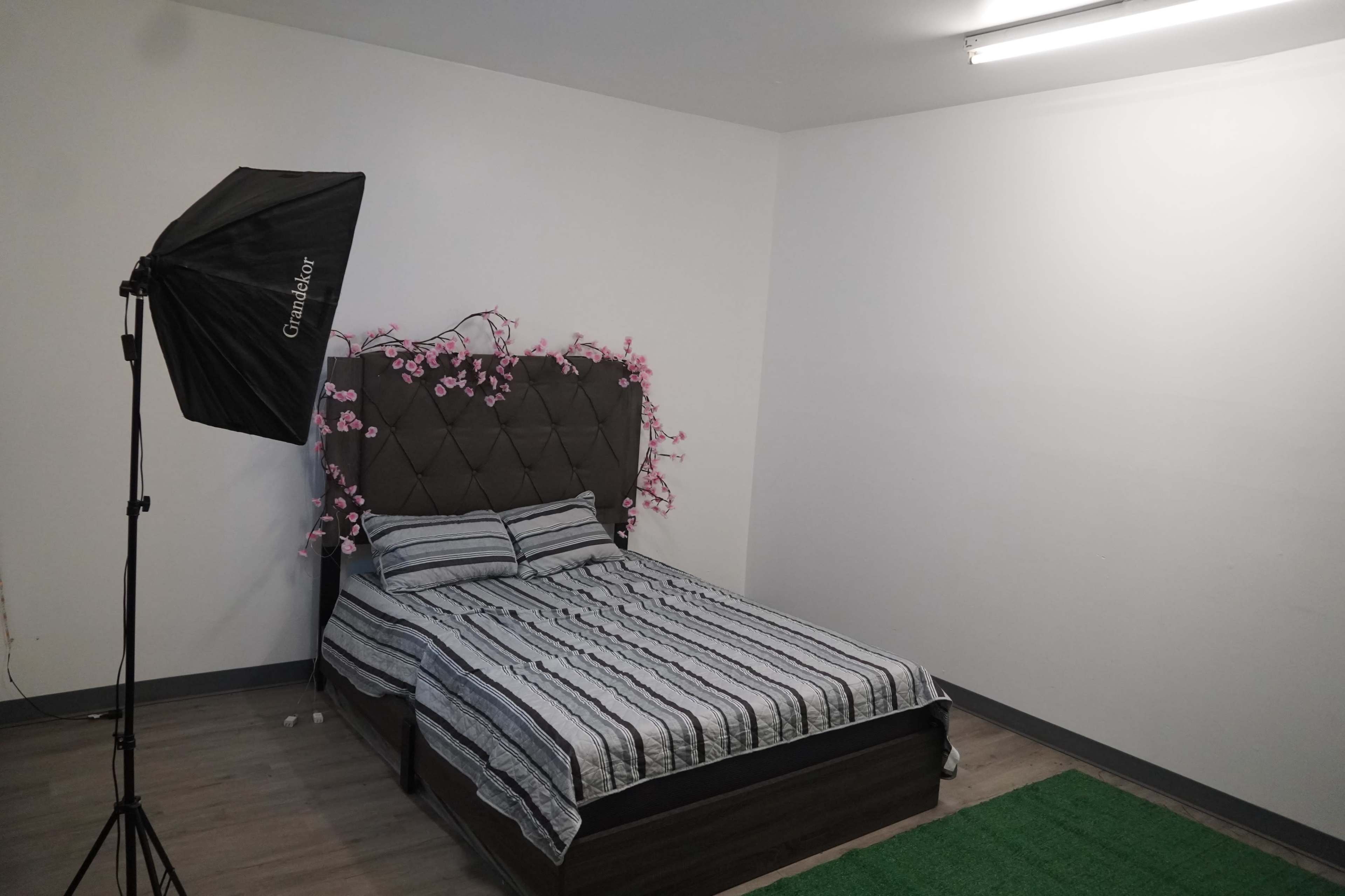 The image shows a bedroom with a bed featuring a striped duvet cover, backdrop of artificial pink flowers, and a photography light stand in the corner.