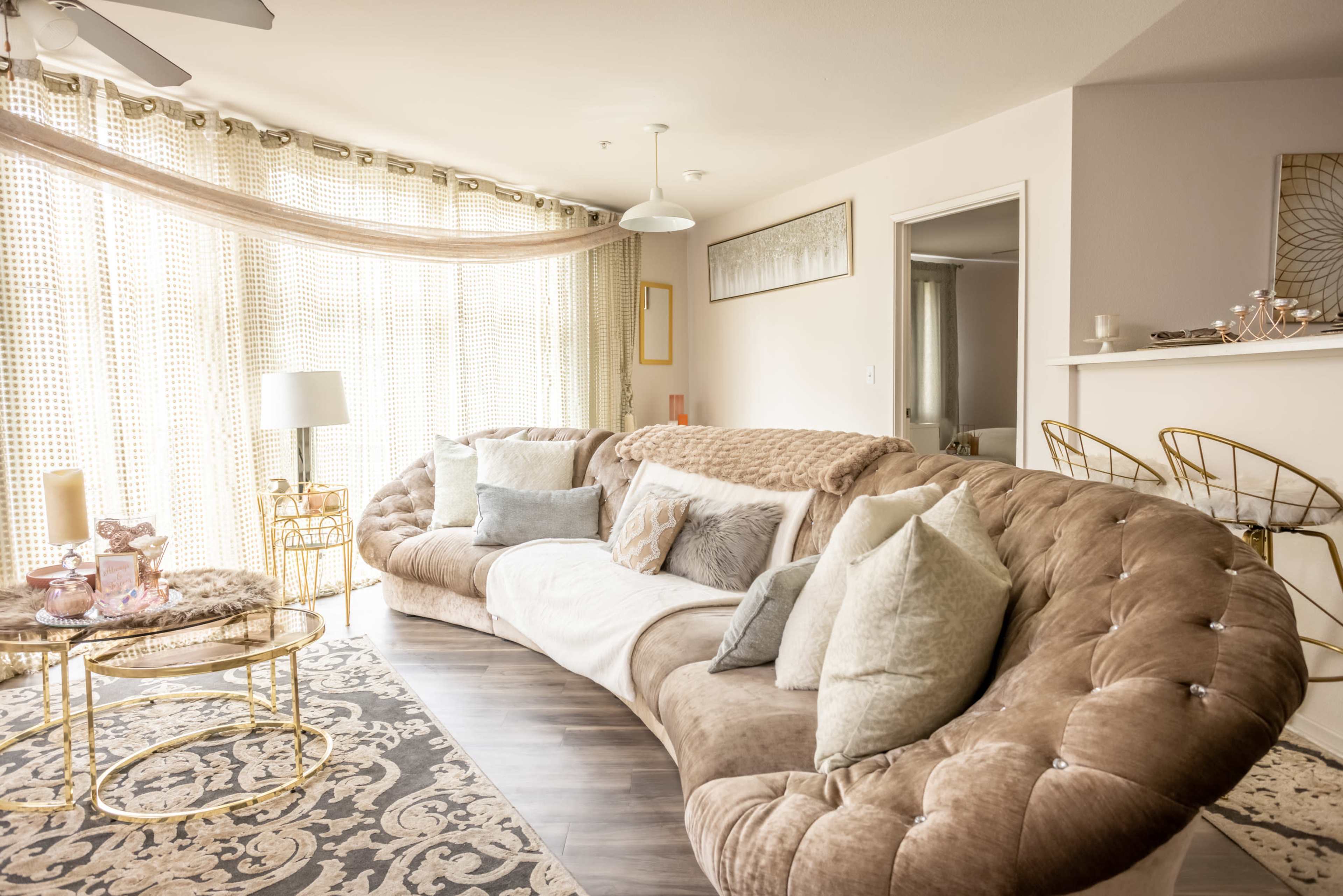 A modern living room features a curved, tufted sofa with decorative pillows, a round coffee table, and large windows adorned with sheer curtains.