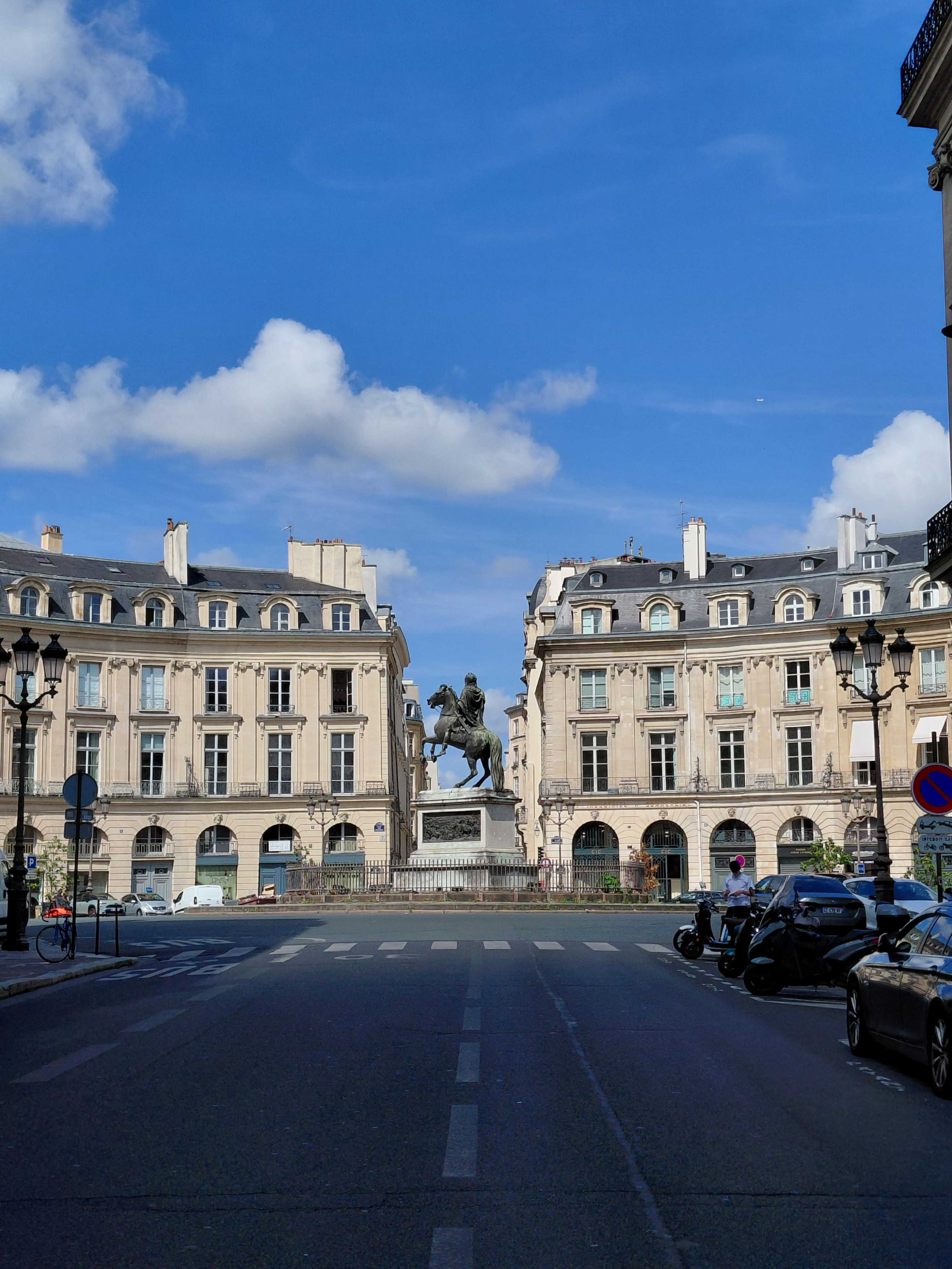 A statue of a horseman stands in the center of a broad street flanked by classical buildings under a blue sky.