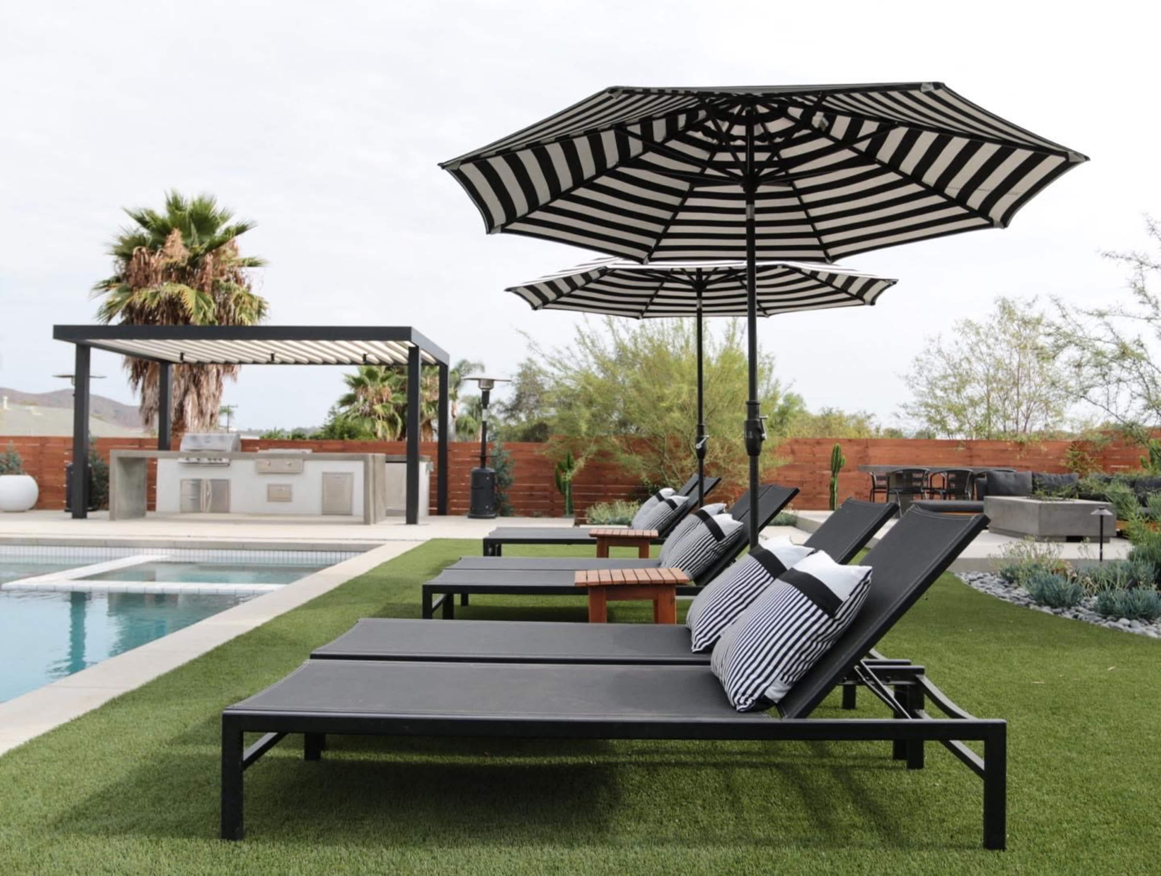 A modern outdoor patio area with poolside lounge chairs under black and white striped umbrellas, featuring a landscaped garden and an outdoor kitchen in the background.