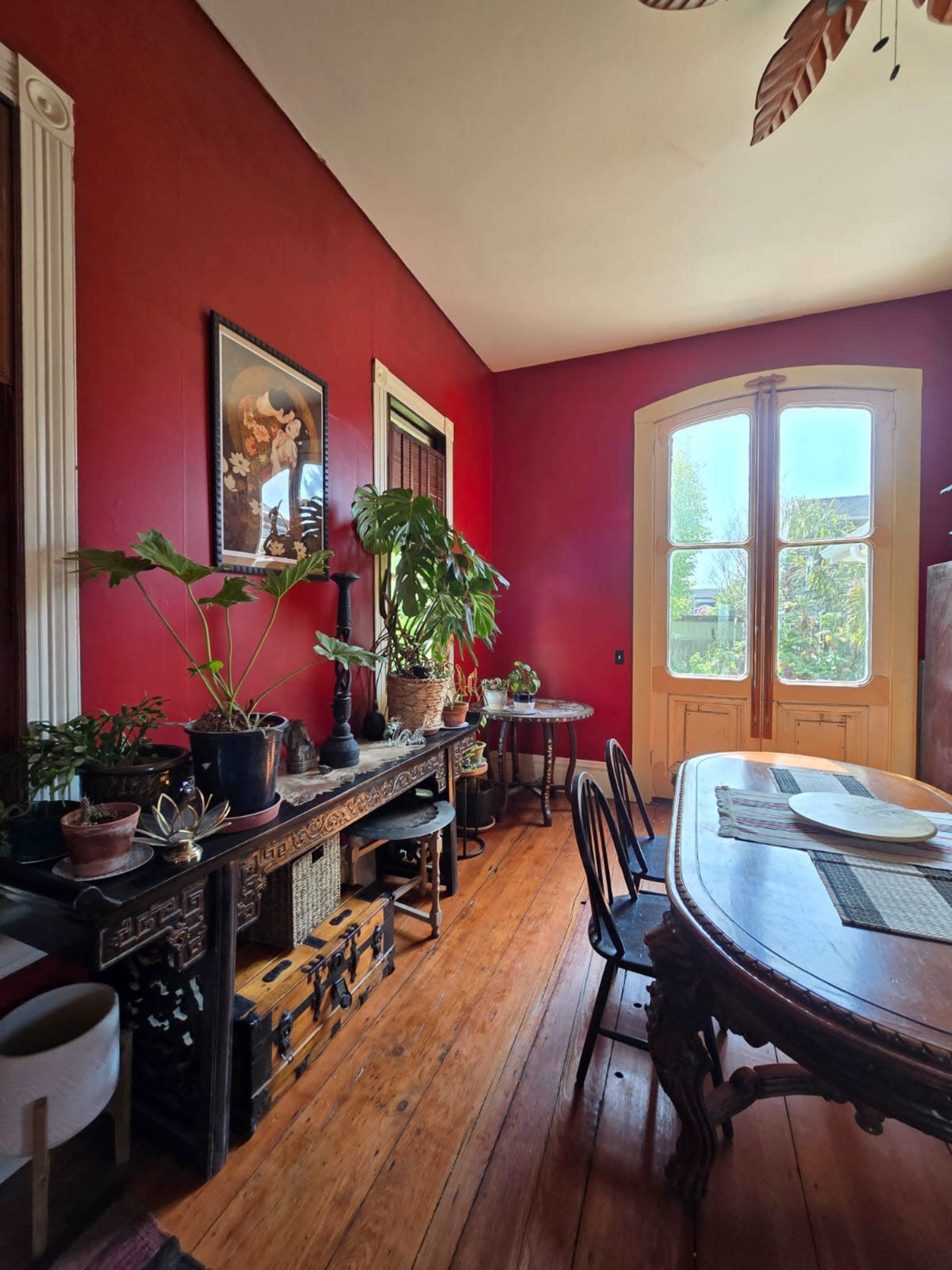 The image depicts a cozy dining area with dark wood furniture, a vibrant red wall, and large windows allowing natural light to enter.