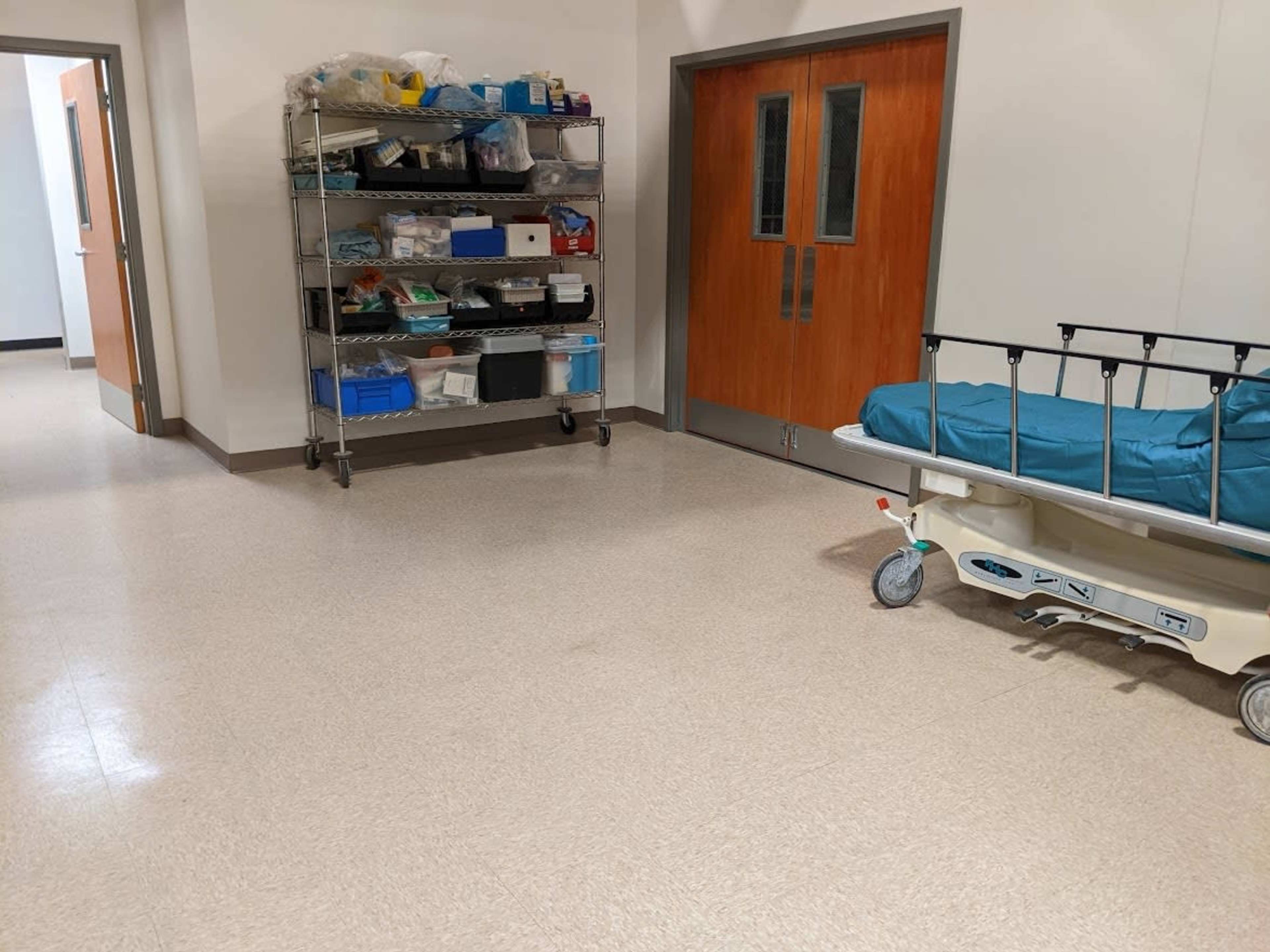 The image shows a hospital corridor with a wheeled hospital bed on one side and a shelving unit filled with medical supplies on the other.