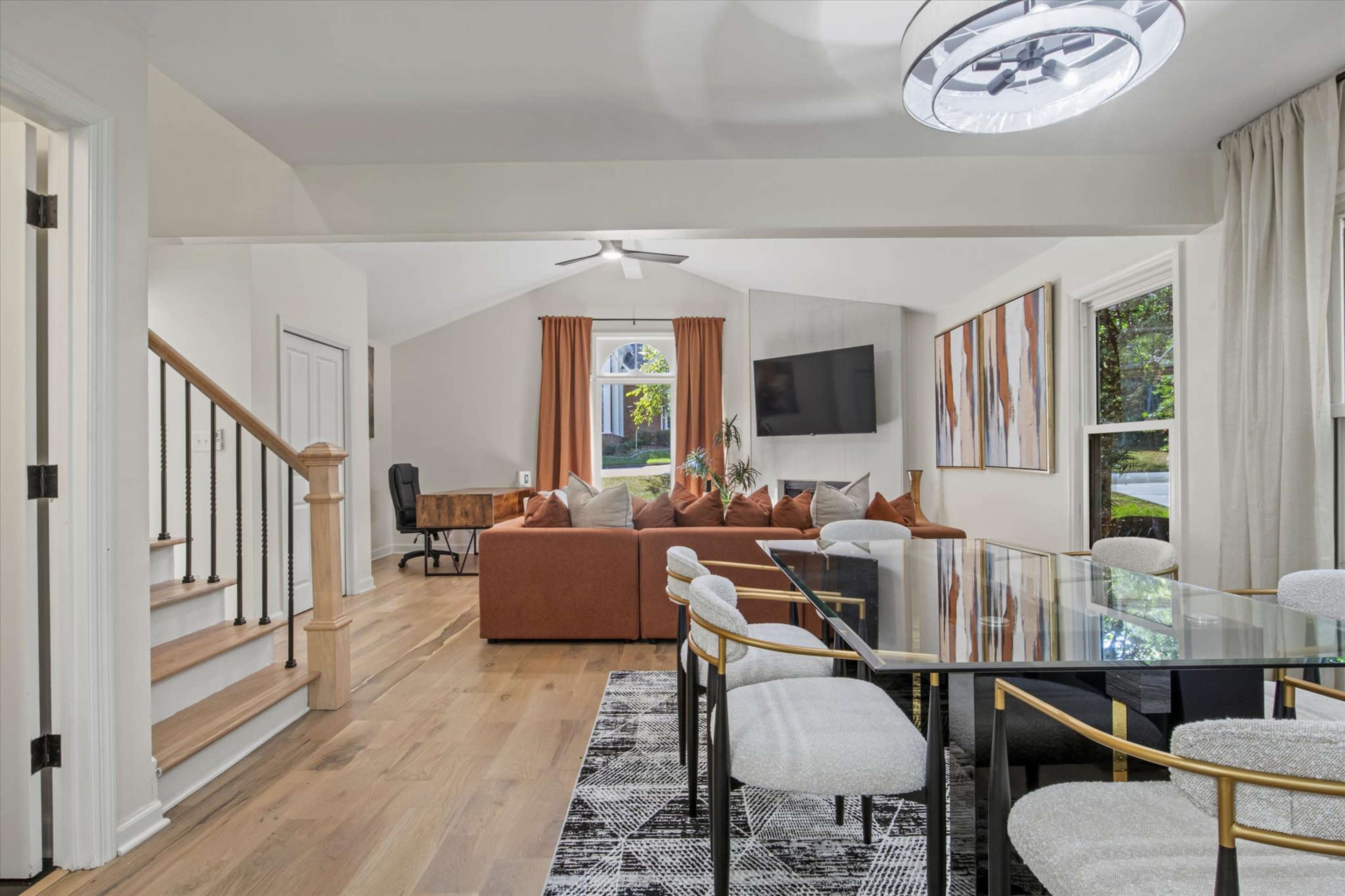 The image shows a modern living area with a comfortable orange sectional sofa, a dining table with upholstered chairs, and large windows allowing natural light to fill the space.
