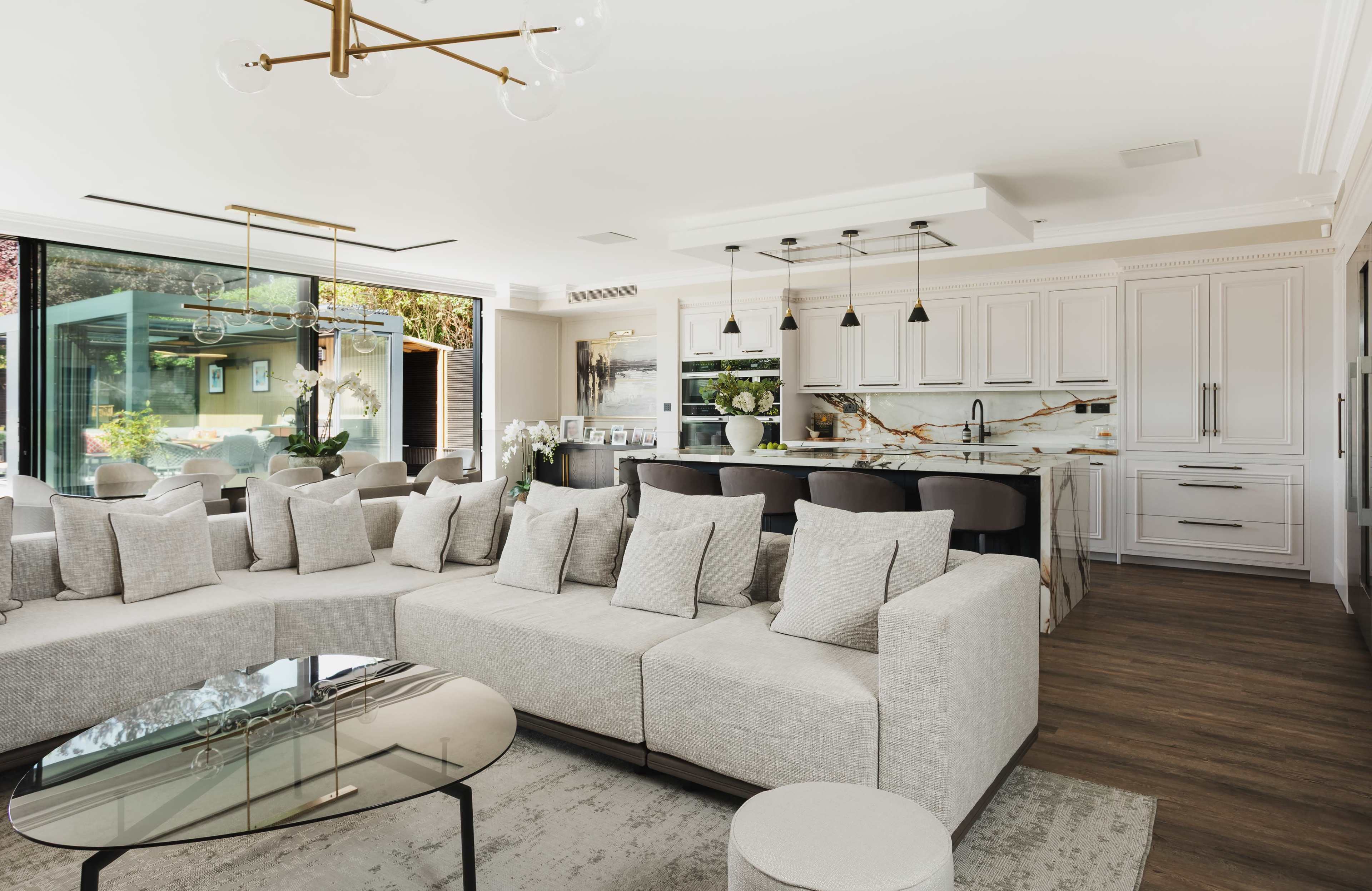 The image shows a modern kitchen and living area featuring a light-colored sectional sofa, a glass coffee table, and a kitchen with white cabinetry and a marble backsplash.