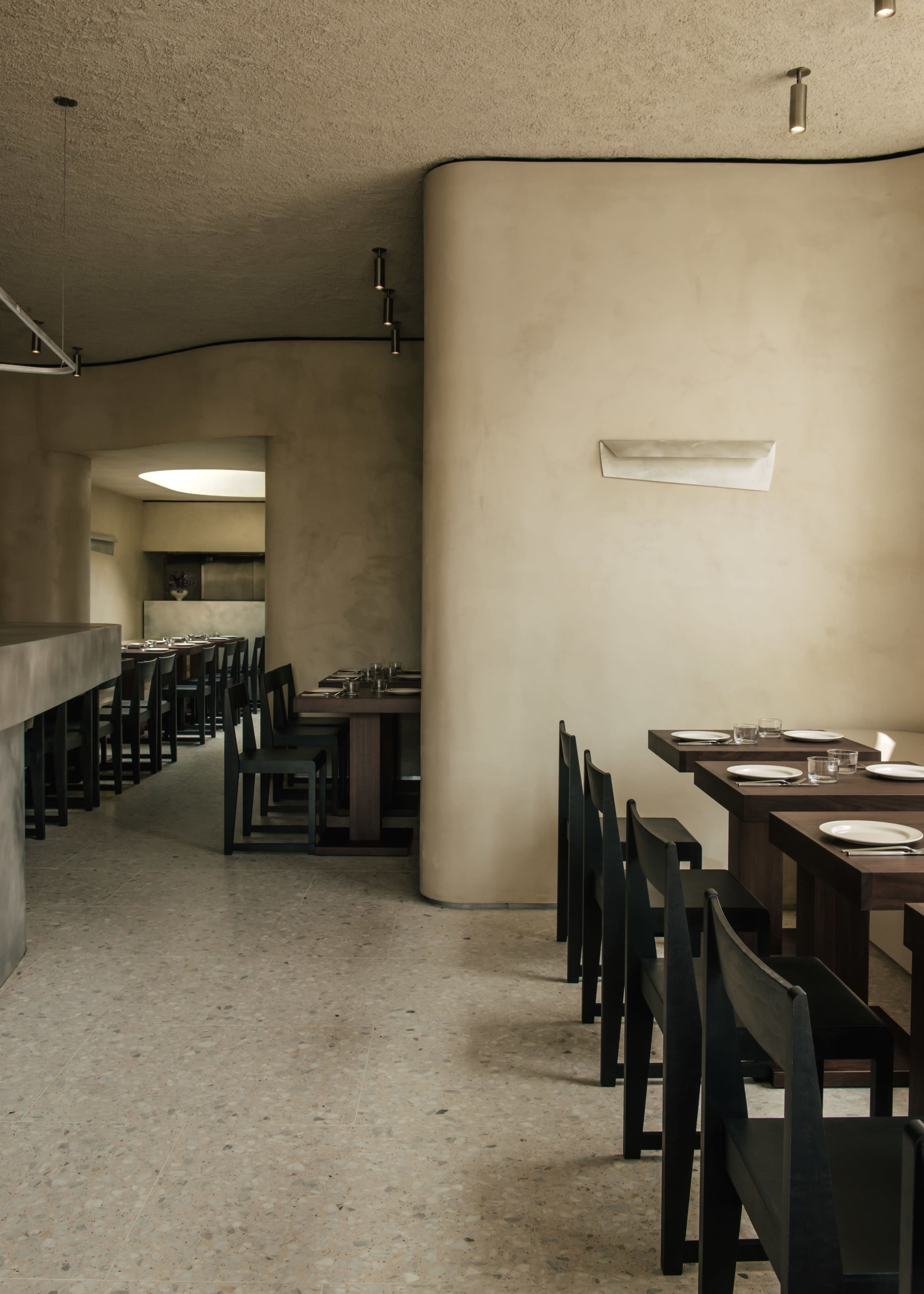 The image shows the interior of a minimalist restaurant with rows of dark wooden tables and chairs against smooth, light-colored walls.