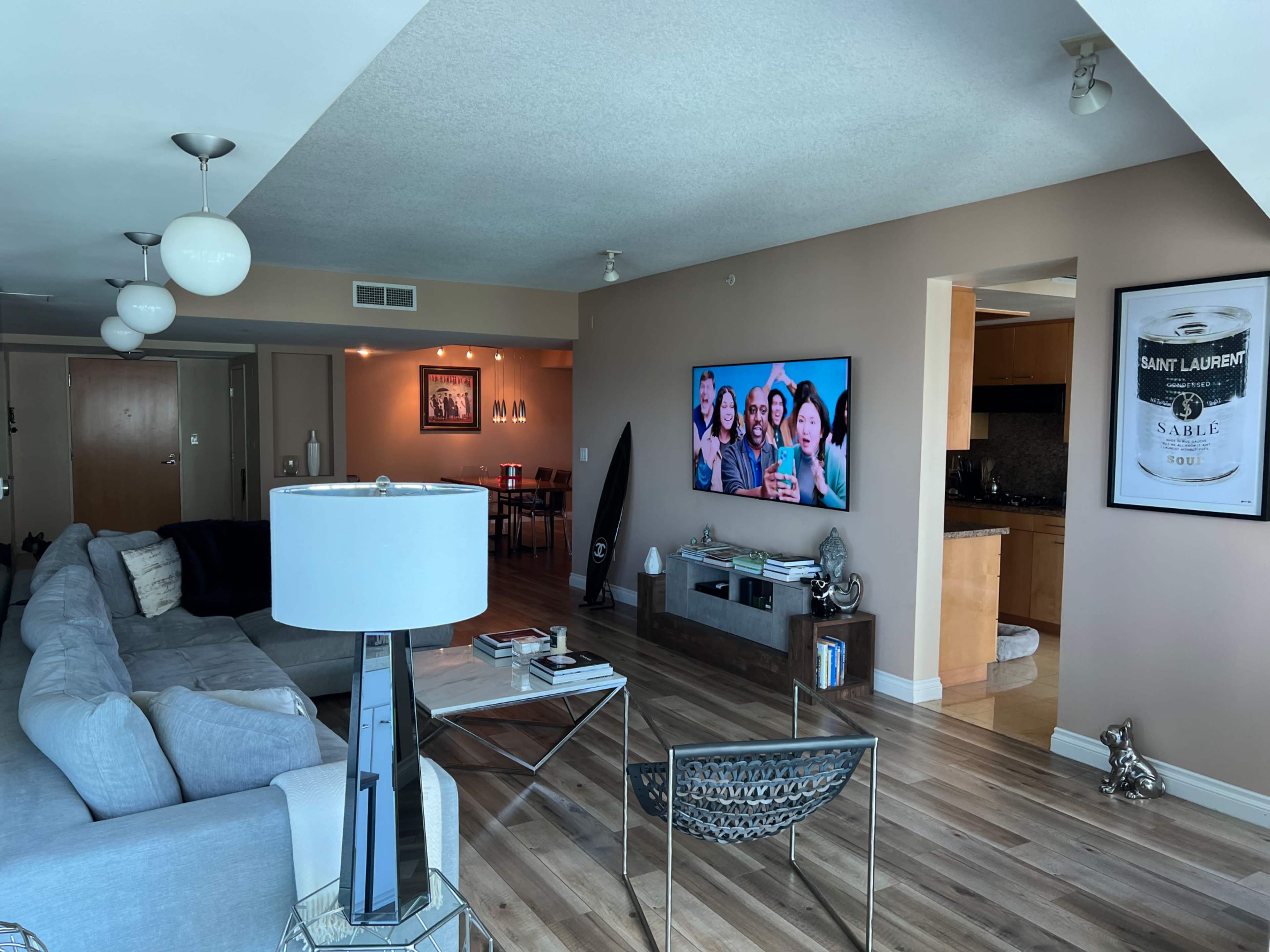 A modern living room with a gray sofa, a large TV displaying a group of people, and a dining area in the background.