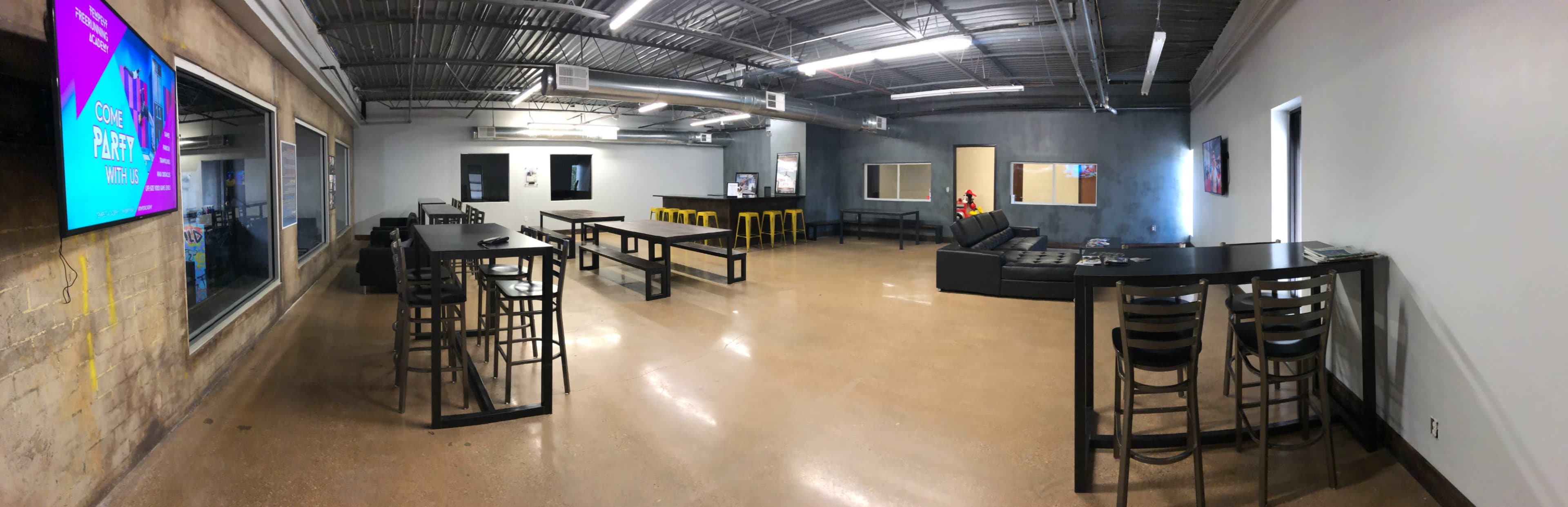 The image shows a spacious indoor area with tables, stools, and a couch, featuring an industrial-style design and multiple entry points.