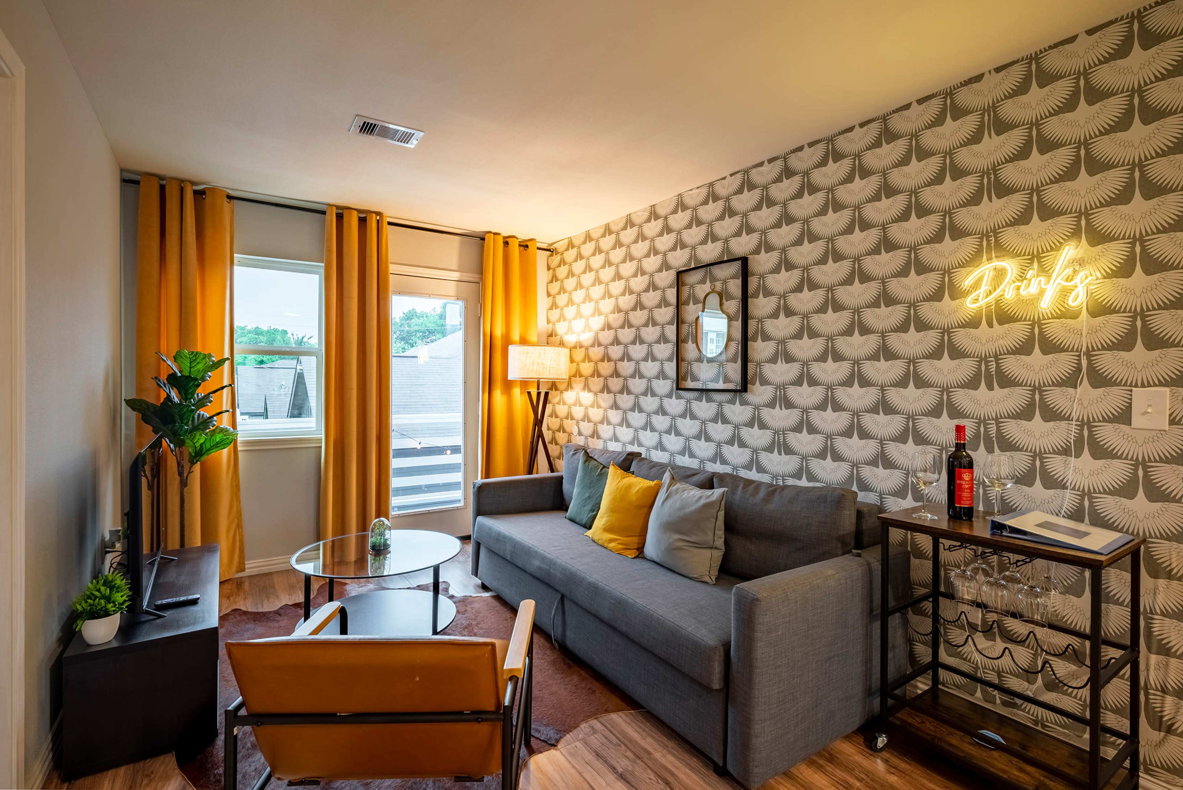 A modern living room features a gray sofa, yellow curtains, a glass coffee table, and a bar cart against a textured wallpaper backdrop.