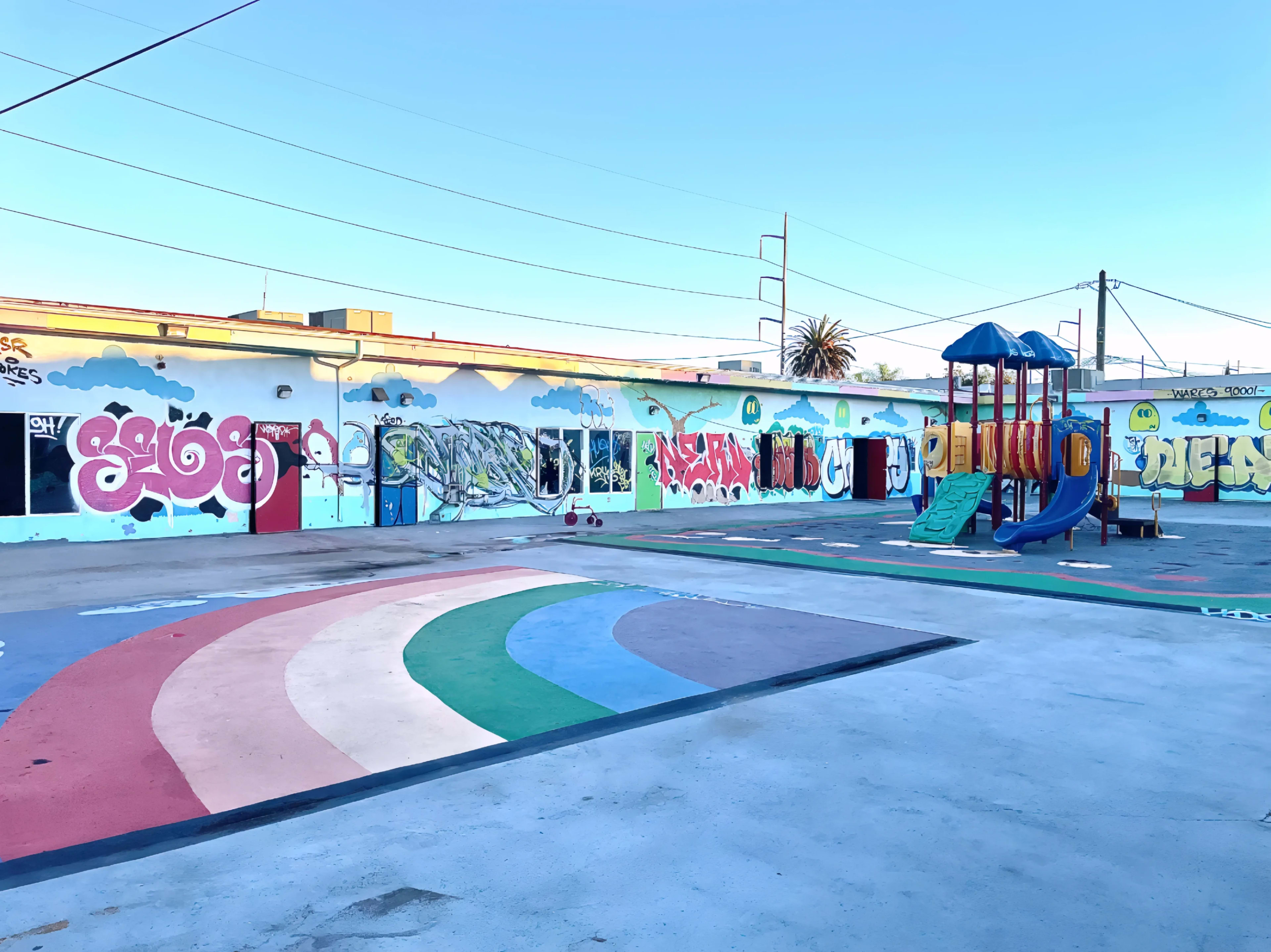 The image shows a playground area with colorful painted pavement and a play structure set against a backdrop of vibrant graffiti-covered walls.