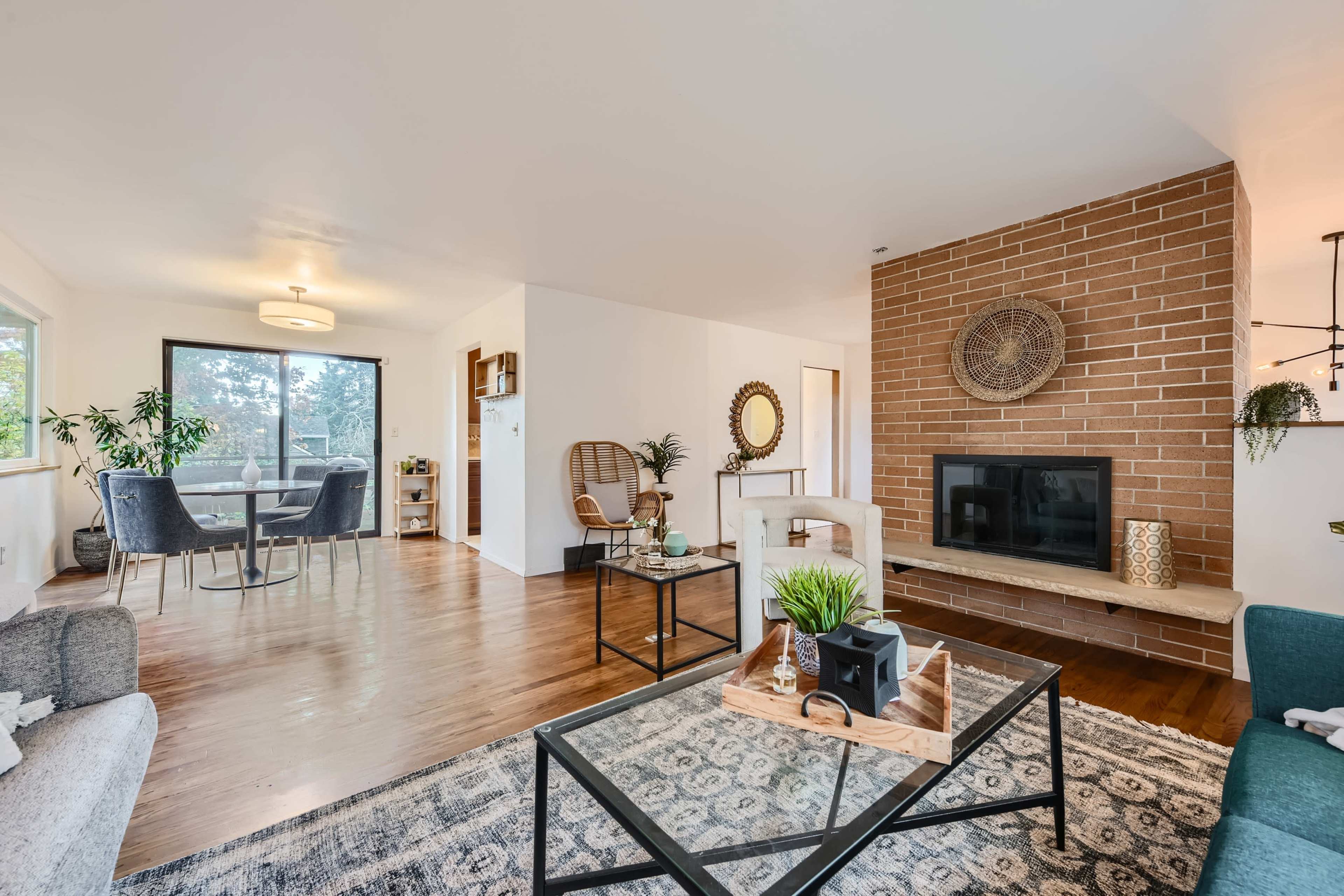 The image shows a modern living room featuring a brown brick fireplace, a glass coffee table, and a dining area with wooden flooring.