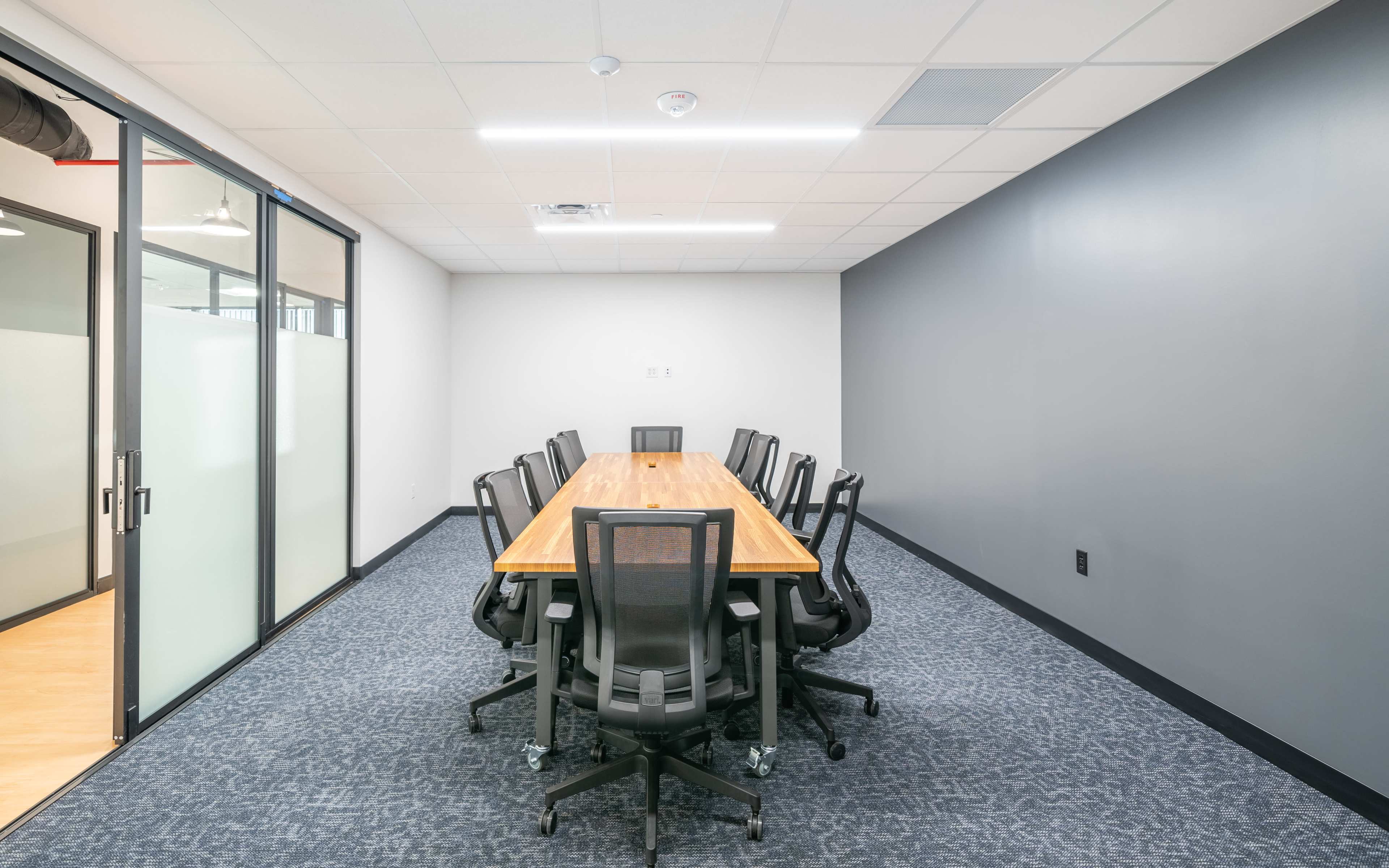 A conference room features a long wooden table surrounded by ten black office chairs, with glass walls and a gray painted wall.