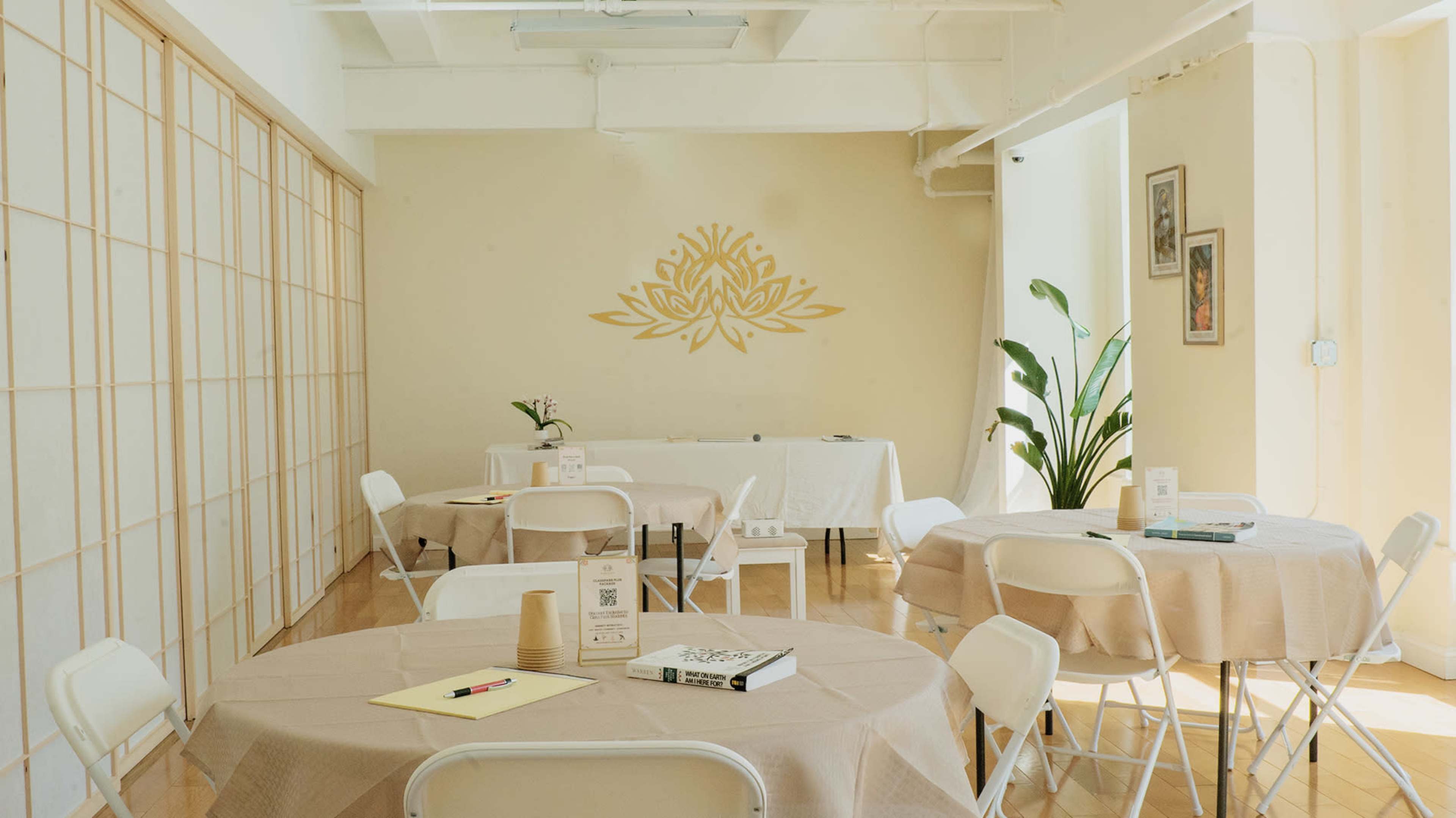 A bright, minimalist room features several round tables covered with beige tablecloths, arranged with chairs, and a decorative wall with a gold lotus design.