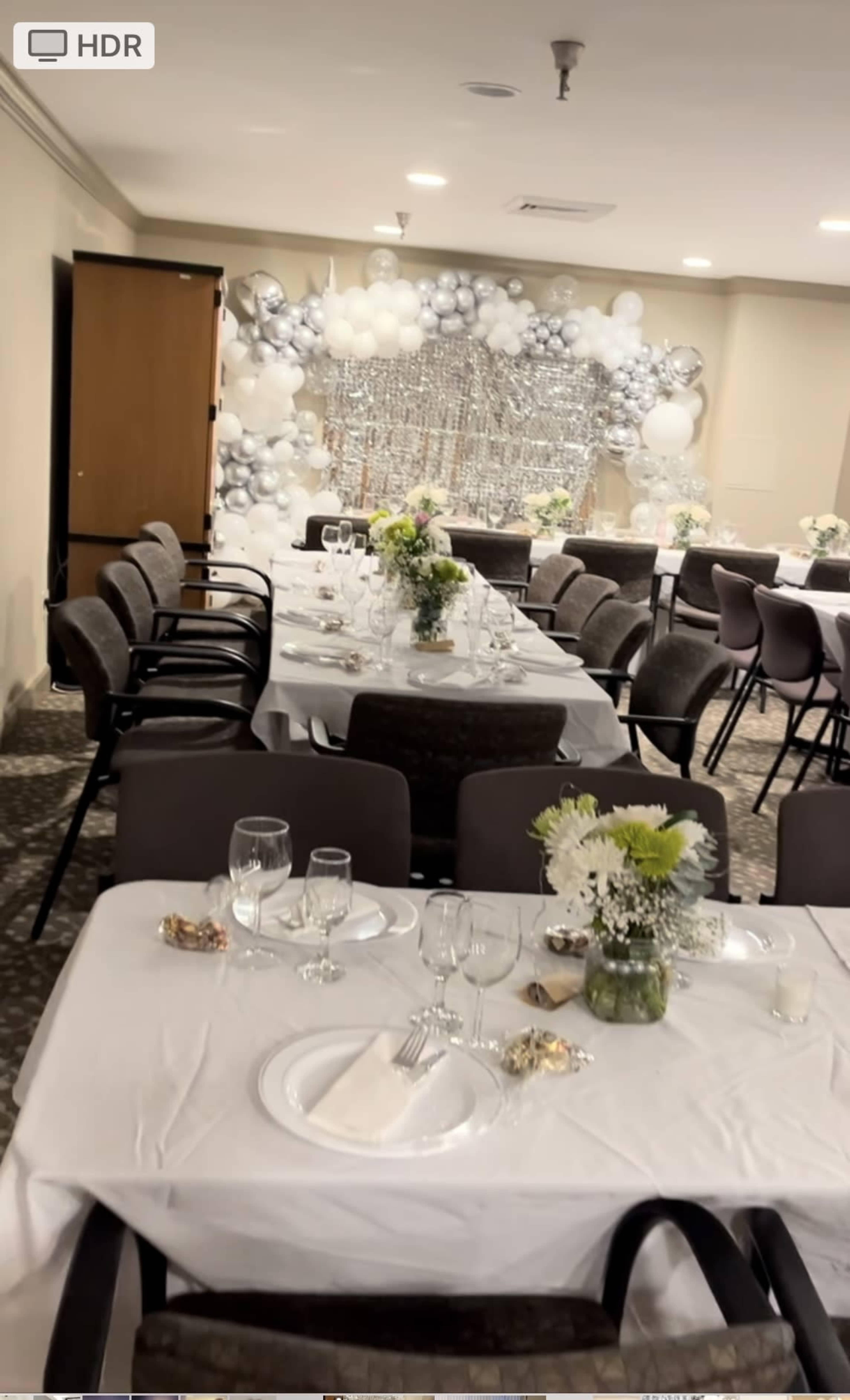 The image shows a banquet hall set for a formal event, featuring white tablecloths, glassware, and a decorative backdrop of balloons and silver accents.