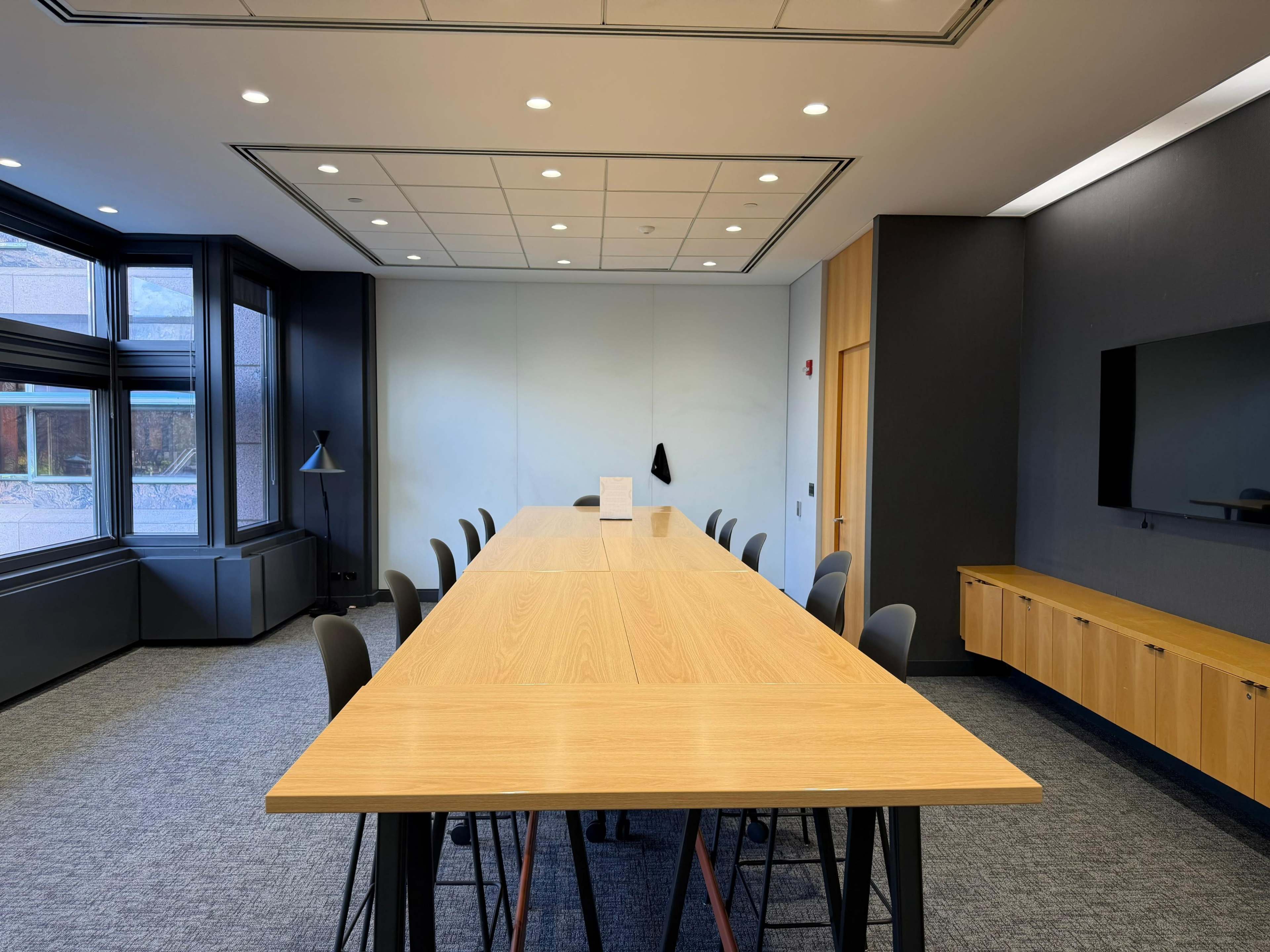 The image shows a large conference room with a long wooden table surrounded by black chairs, featuring large windows and a wall-mounted screen.