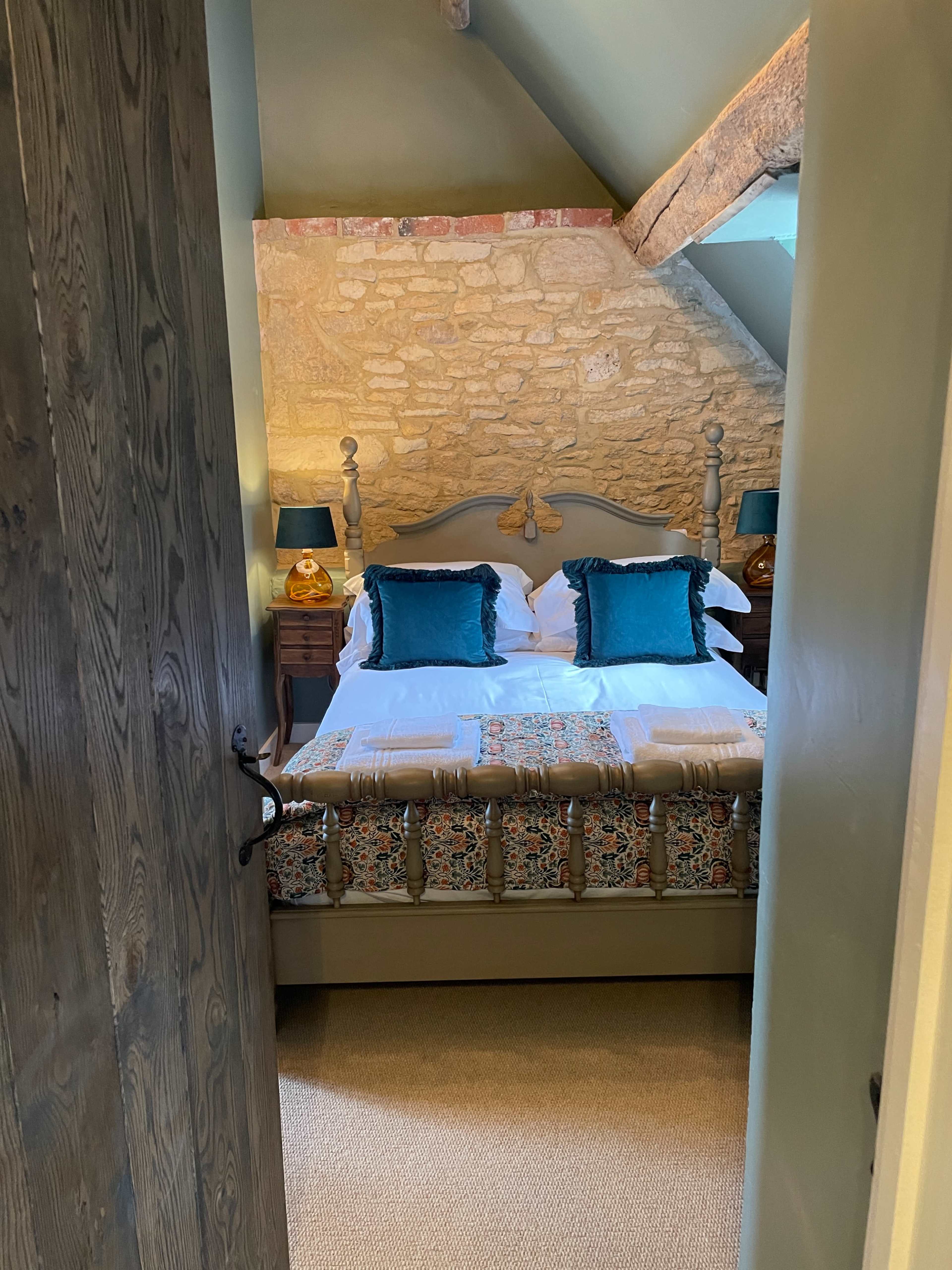 A bedroom featuring a stone wall, a bed with decorative pillows, and two bedside lamps, as viewed through an open door.