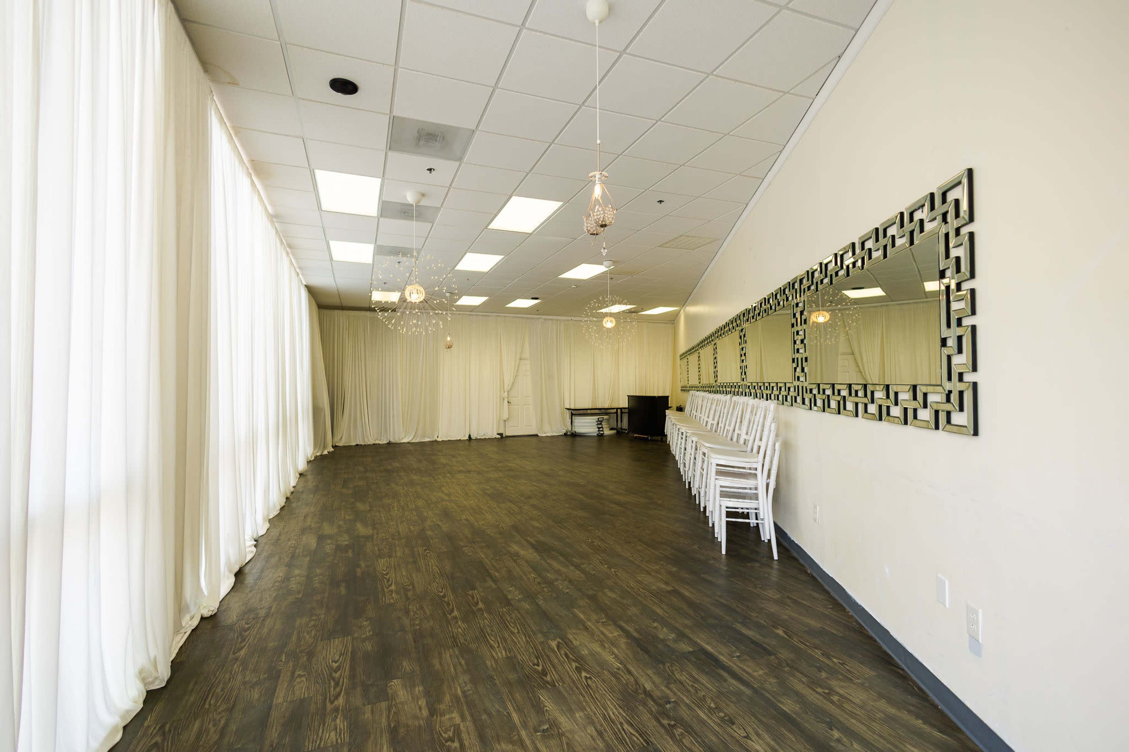 The image shows a spacious, empty indoor room with light-colored walls, large windows dressed in white drapes, and a row of white chairs lined against one wall.