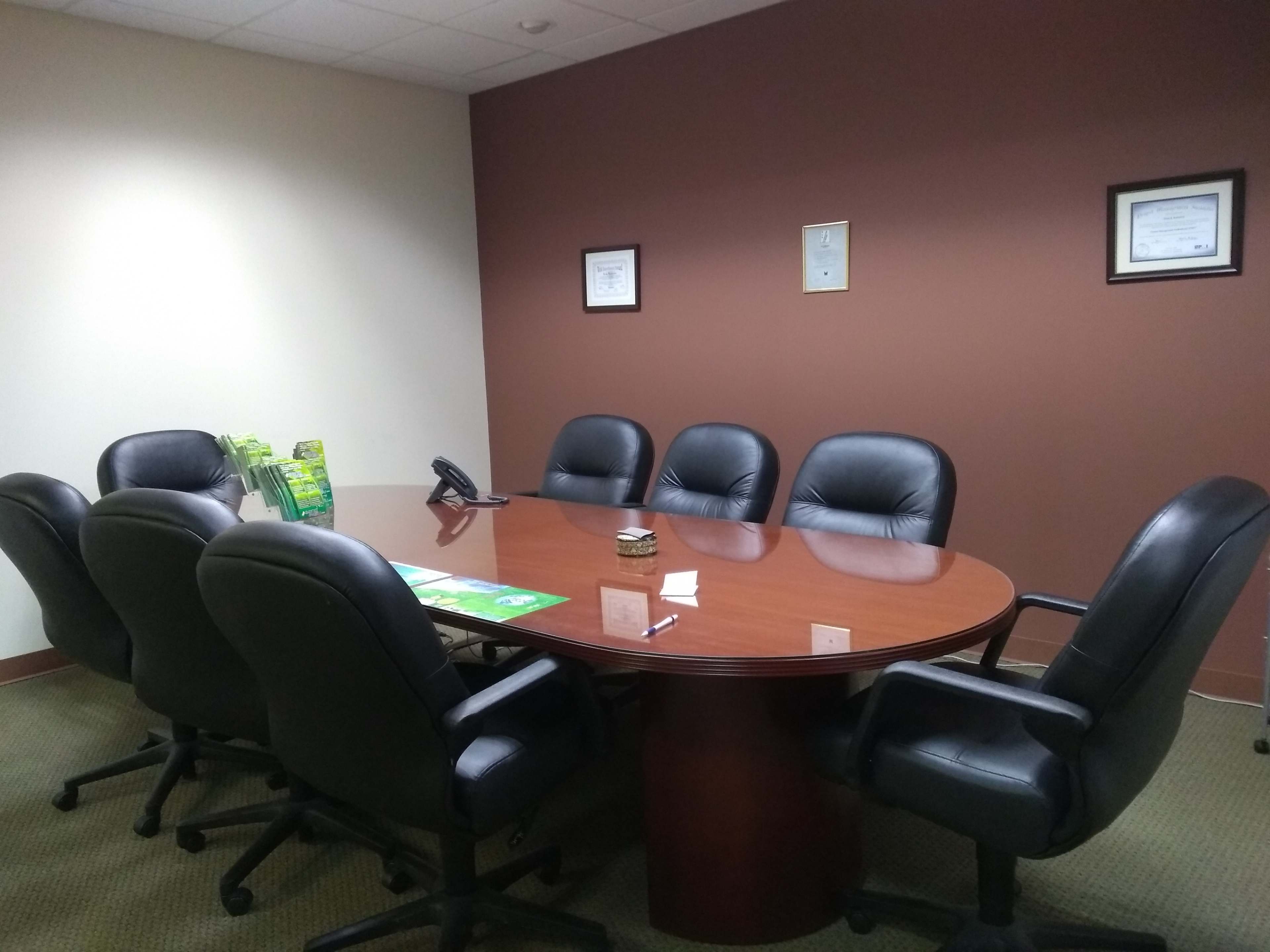 A conference room features a round wooden table surrounded by six black leather chairs, with a phone and materials on the table and certificates displayed on the wall.
