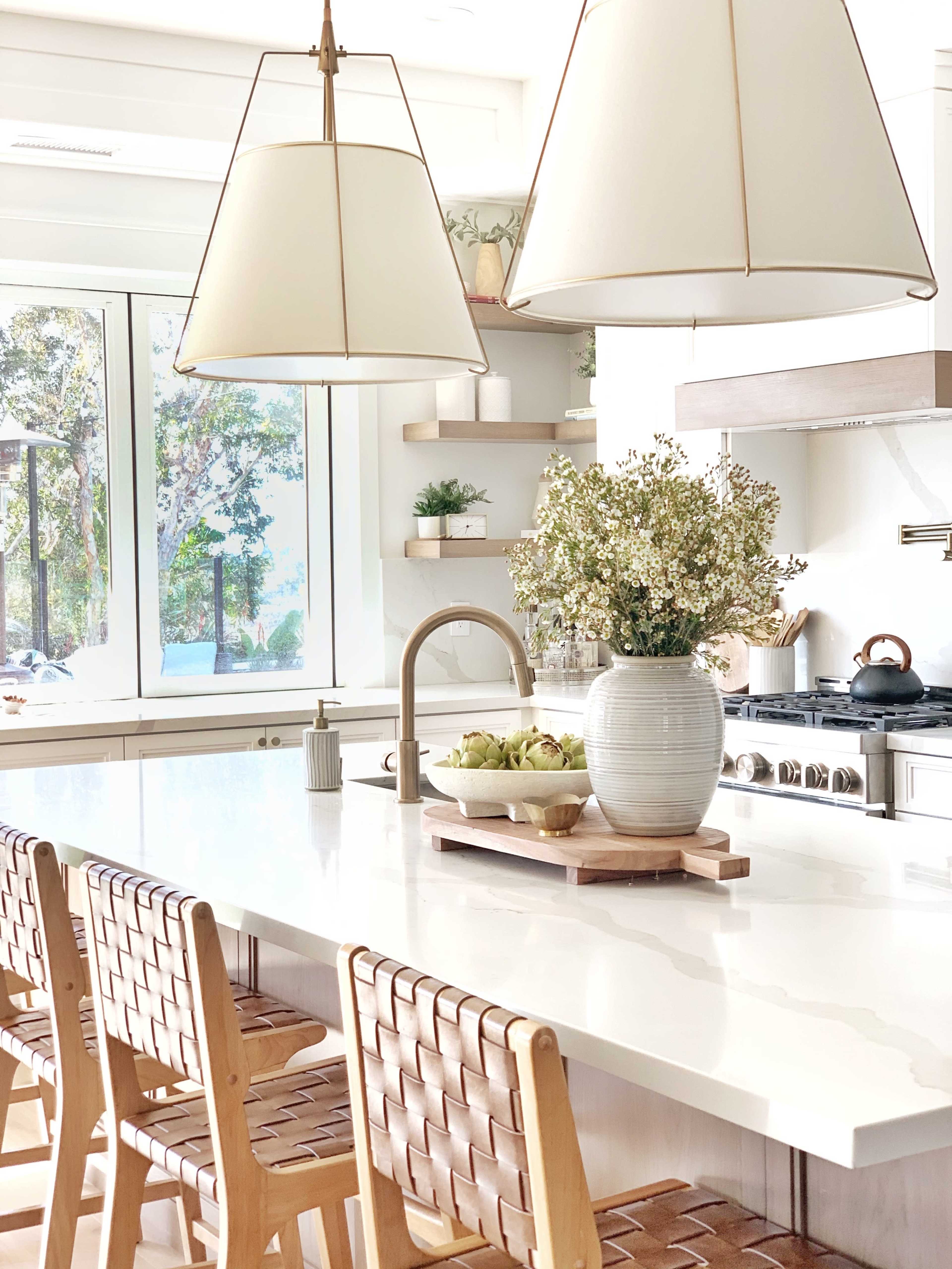 A modern kitchen features a large white countertop, wooden bar stools, and pendant lighting over a centerpiece vase with flowers.