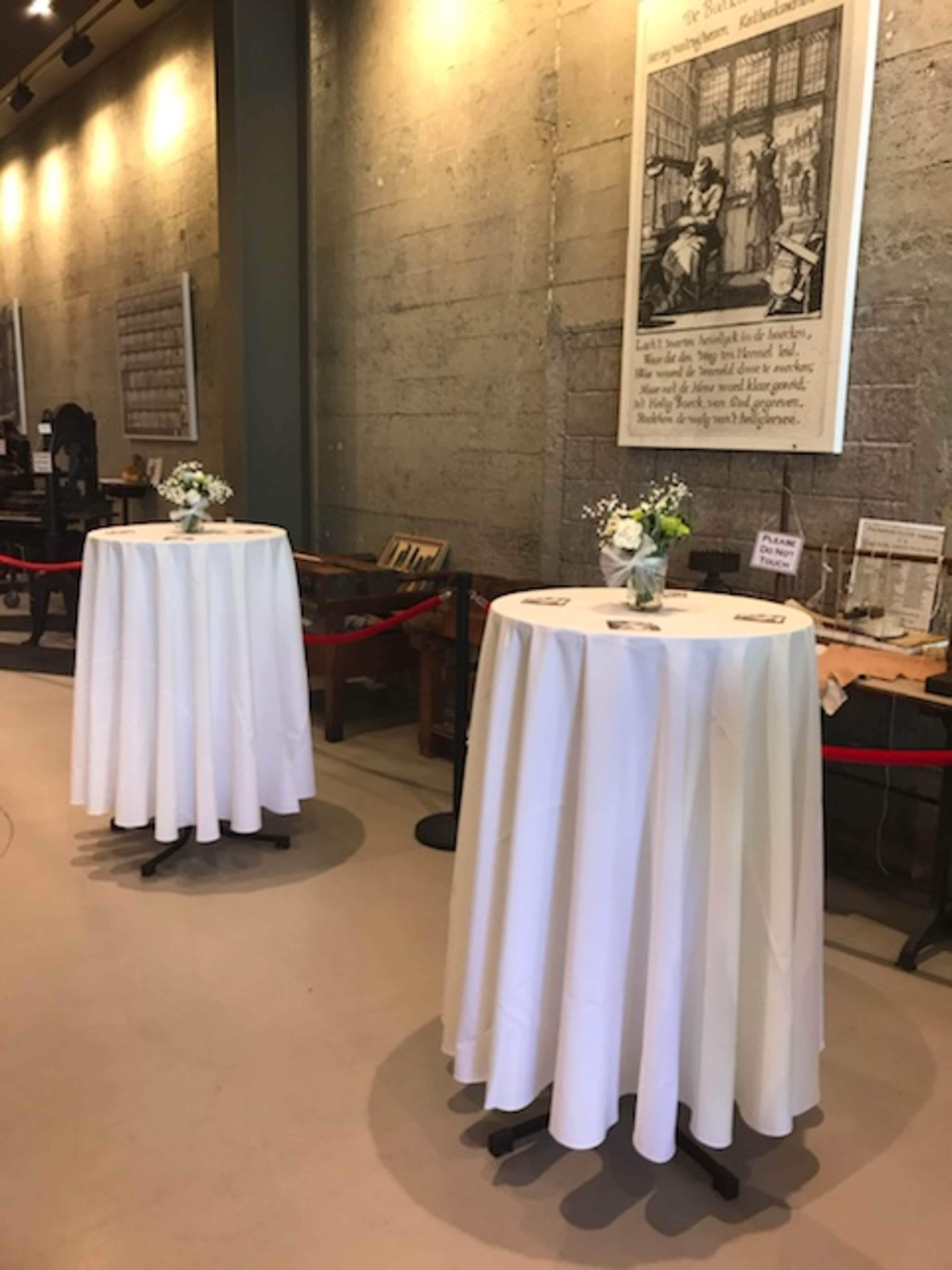 Two round tables covered with white tablecloths, set up in a well-lit space with a concrete wall and a large framed artwork in the background.