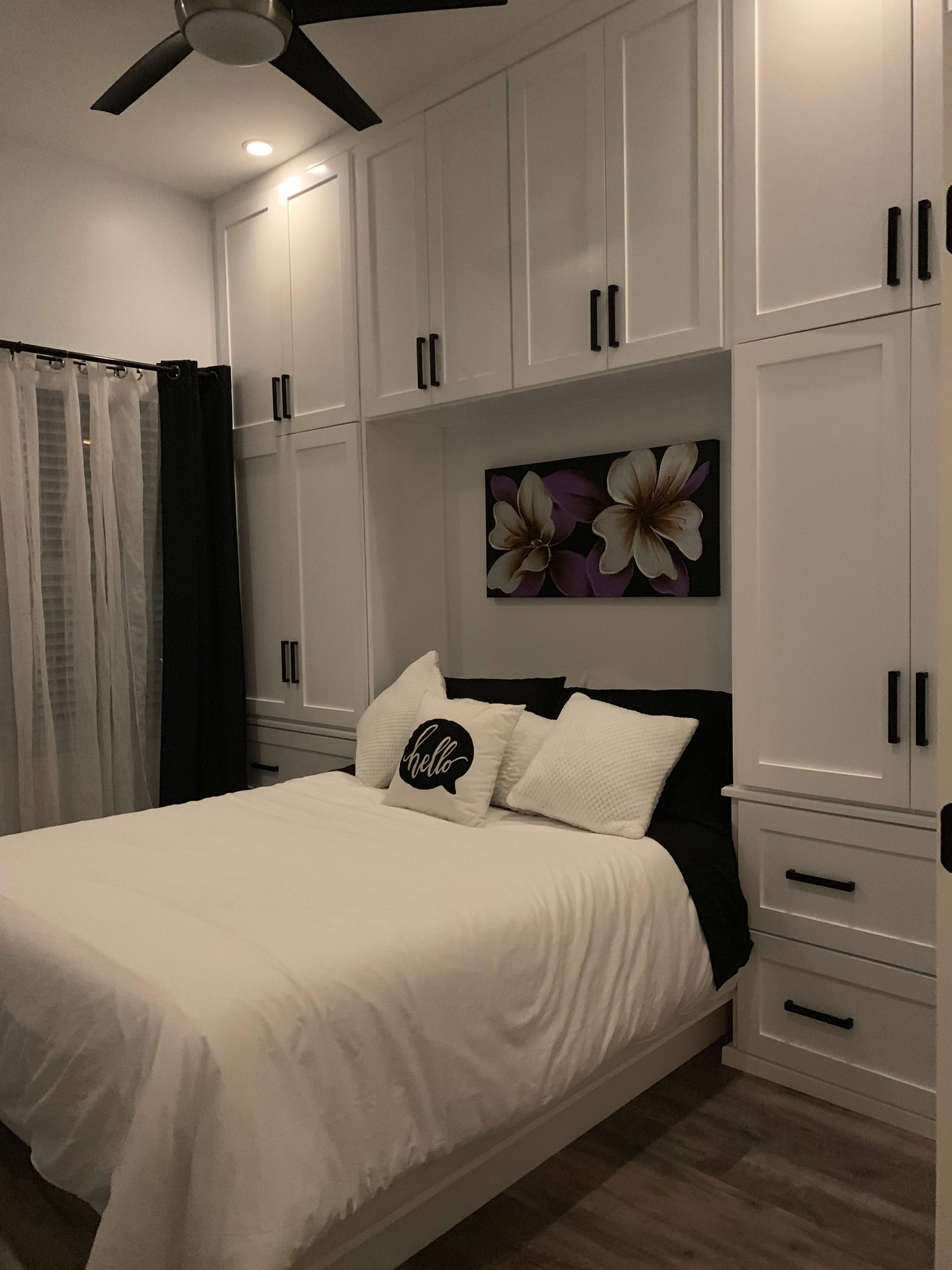 The image shows a small bedroom with a bed featuring white bedding and decorative pillows against a backdrop of built-in white cabinetry and a floral artwork.