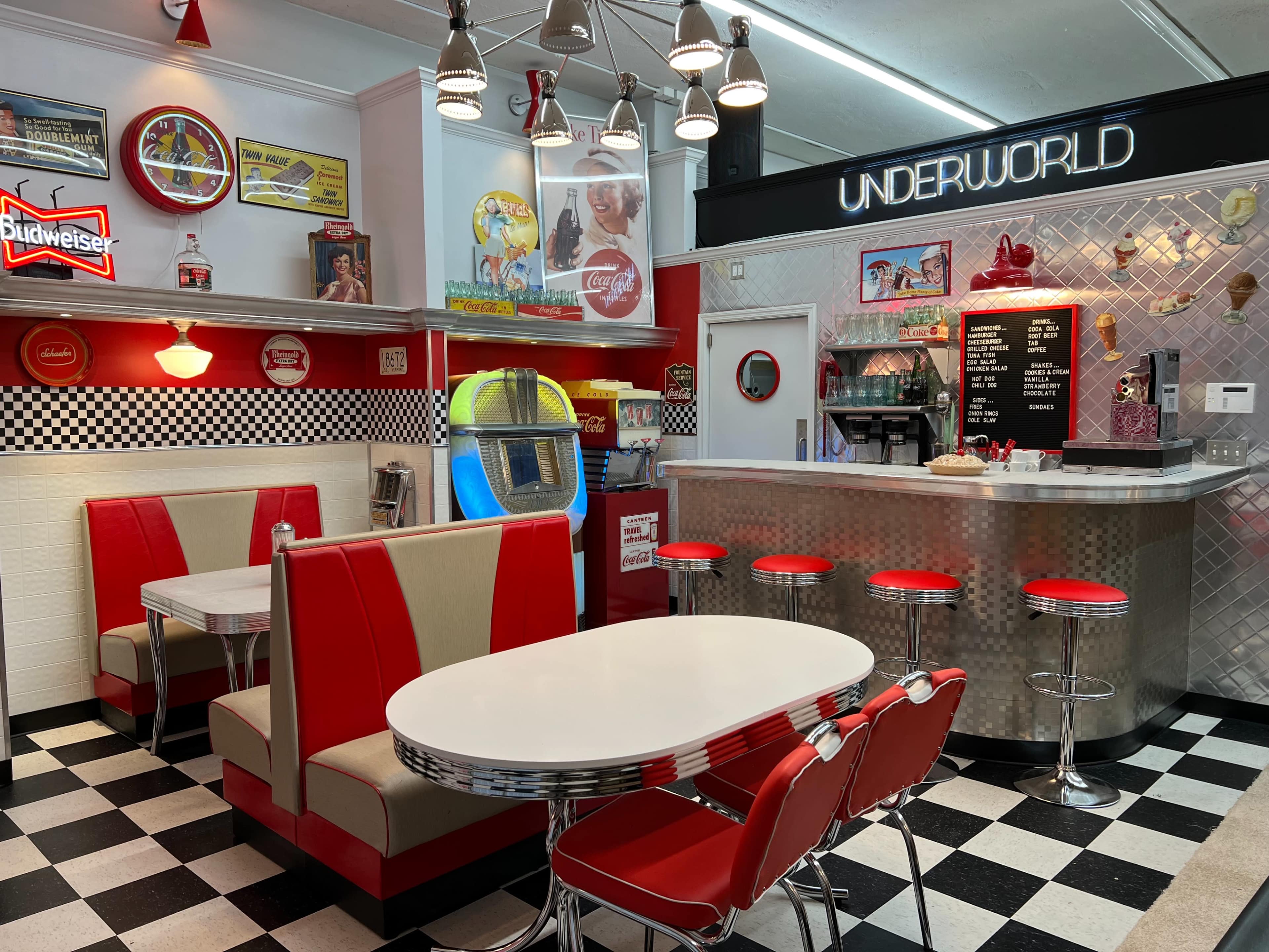 A retro diner interior featuring red and white booths, a checkered floor, a jukebox, and a stainless steel counter with stools.