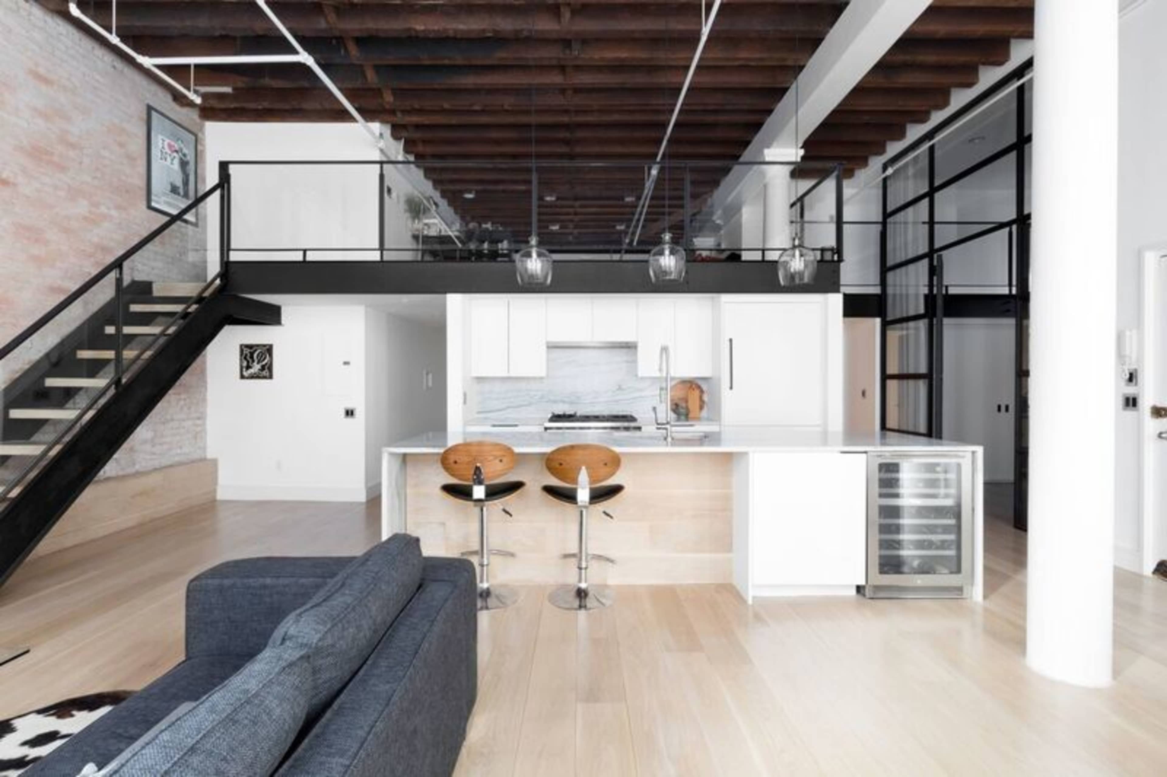 A modern kitchen with a central island and two bar stools, adjacent to a living area featuring a gray sofa and a staircase leading to a lofted space.