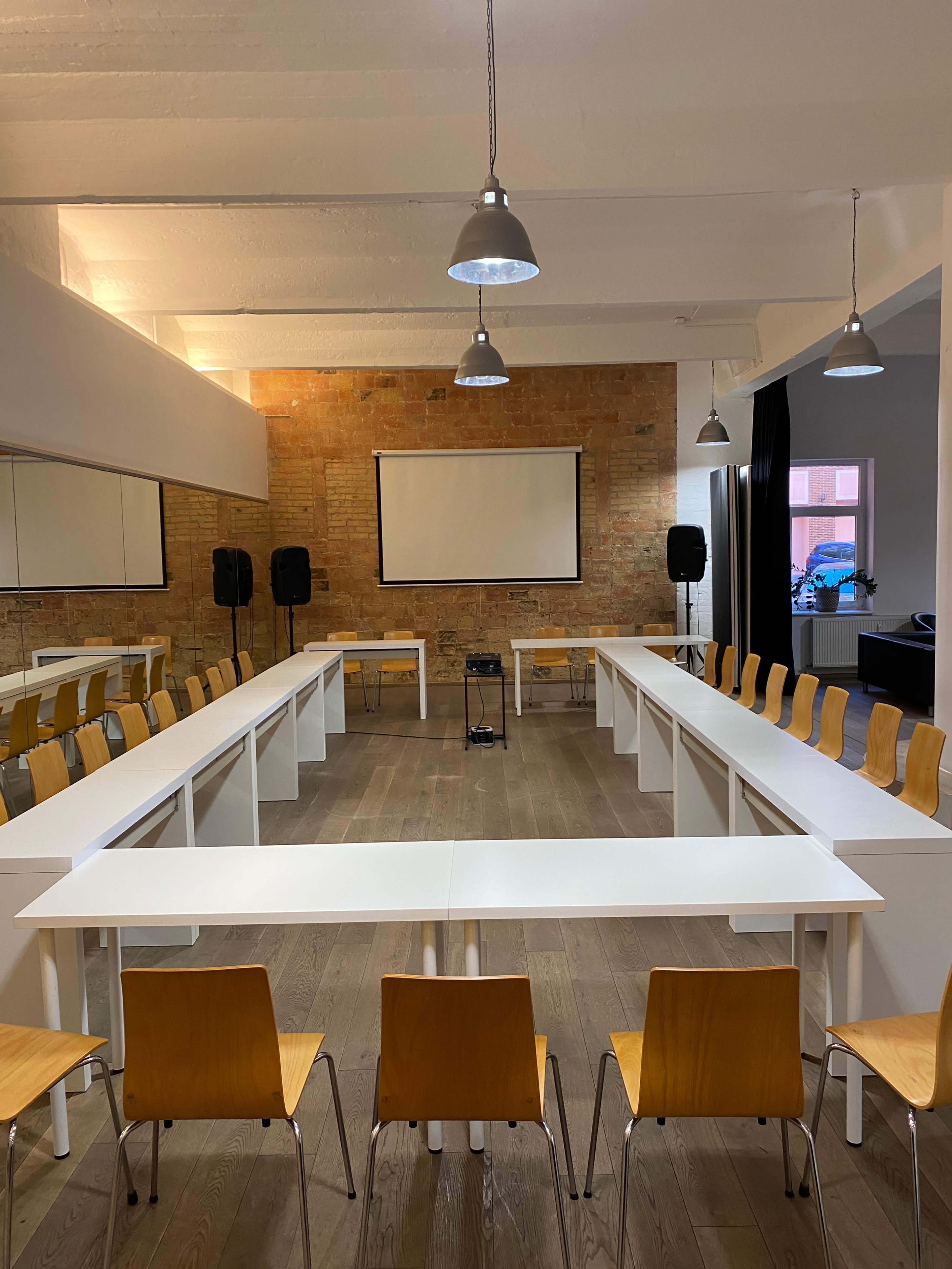 A spacious meeting room features long white tables arranged in a rectangle with wooden chairs, a projector screen at the front, and exposed brick walls.