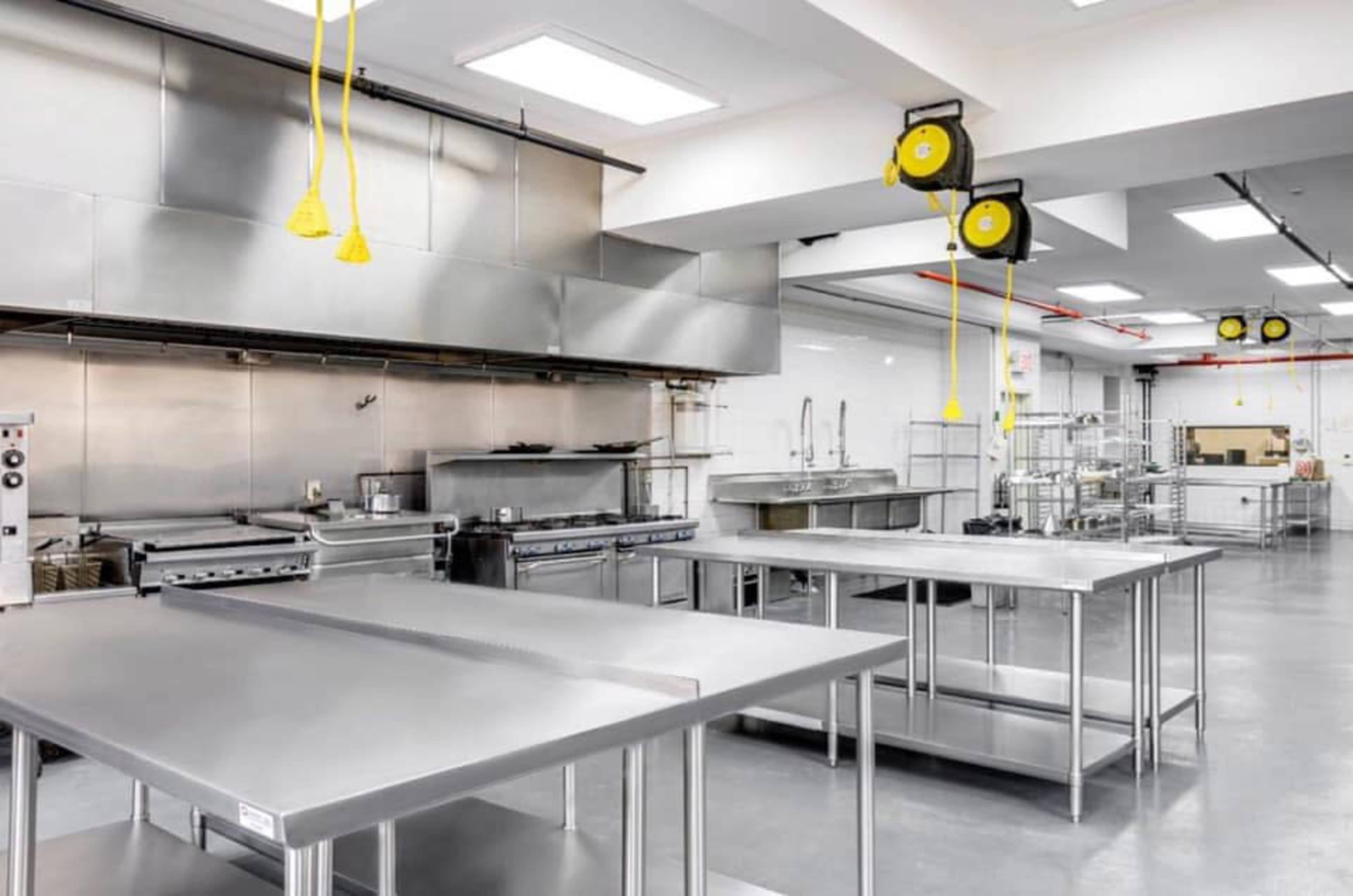 The image shows a spacious, commercial kitchen equipped with stainless steel tables, cooking appliances, and overhead yellow power cords.
