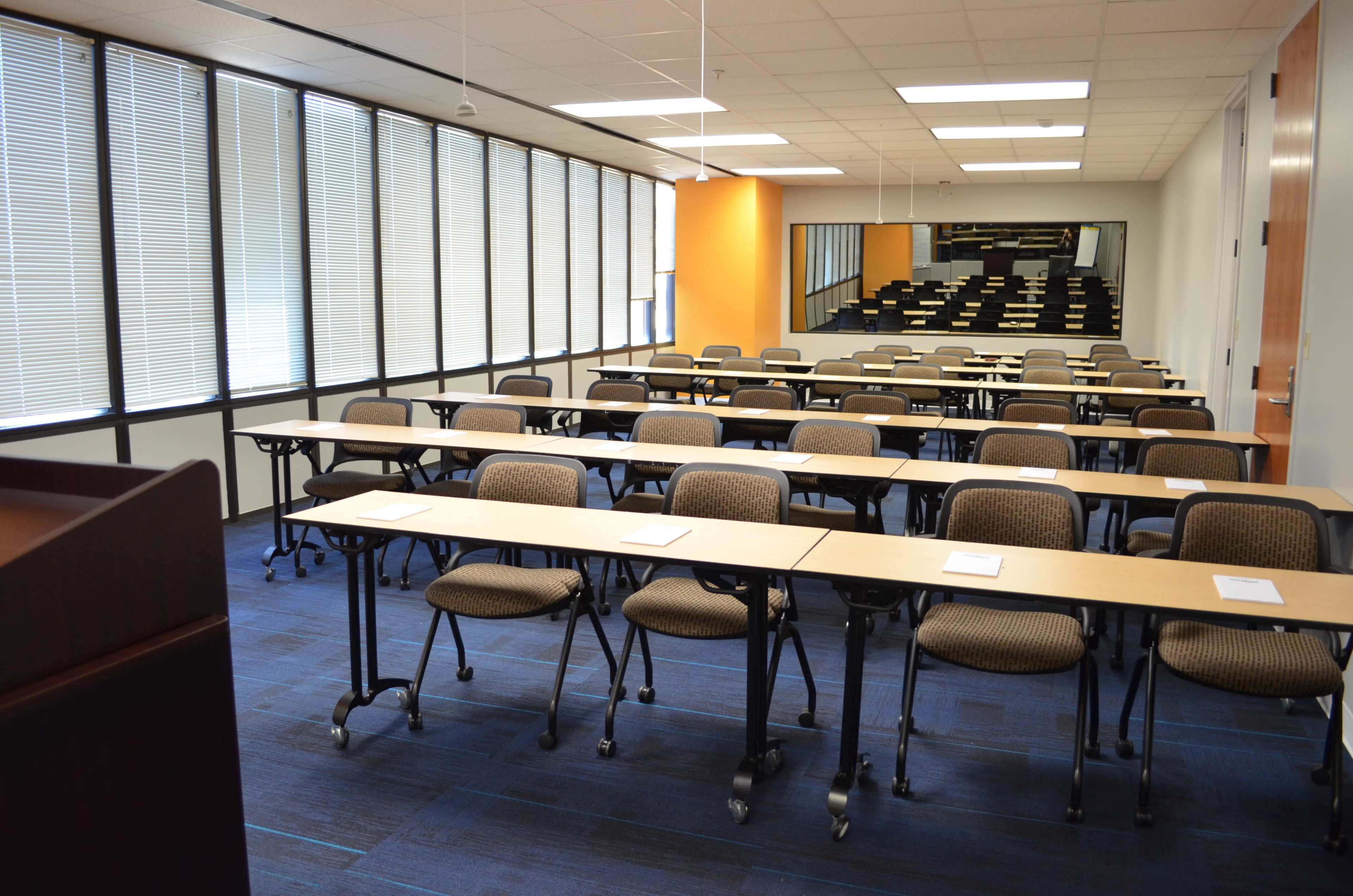 A classroom with rows of tables and chairs facing a presentation area, featuring large windows on one side and a staircase leading to an elevated section.