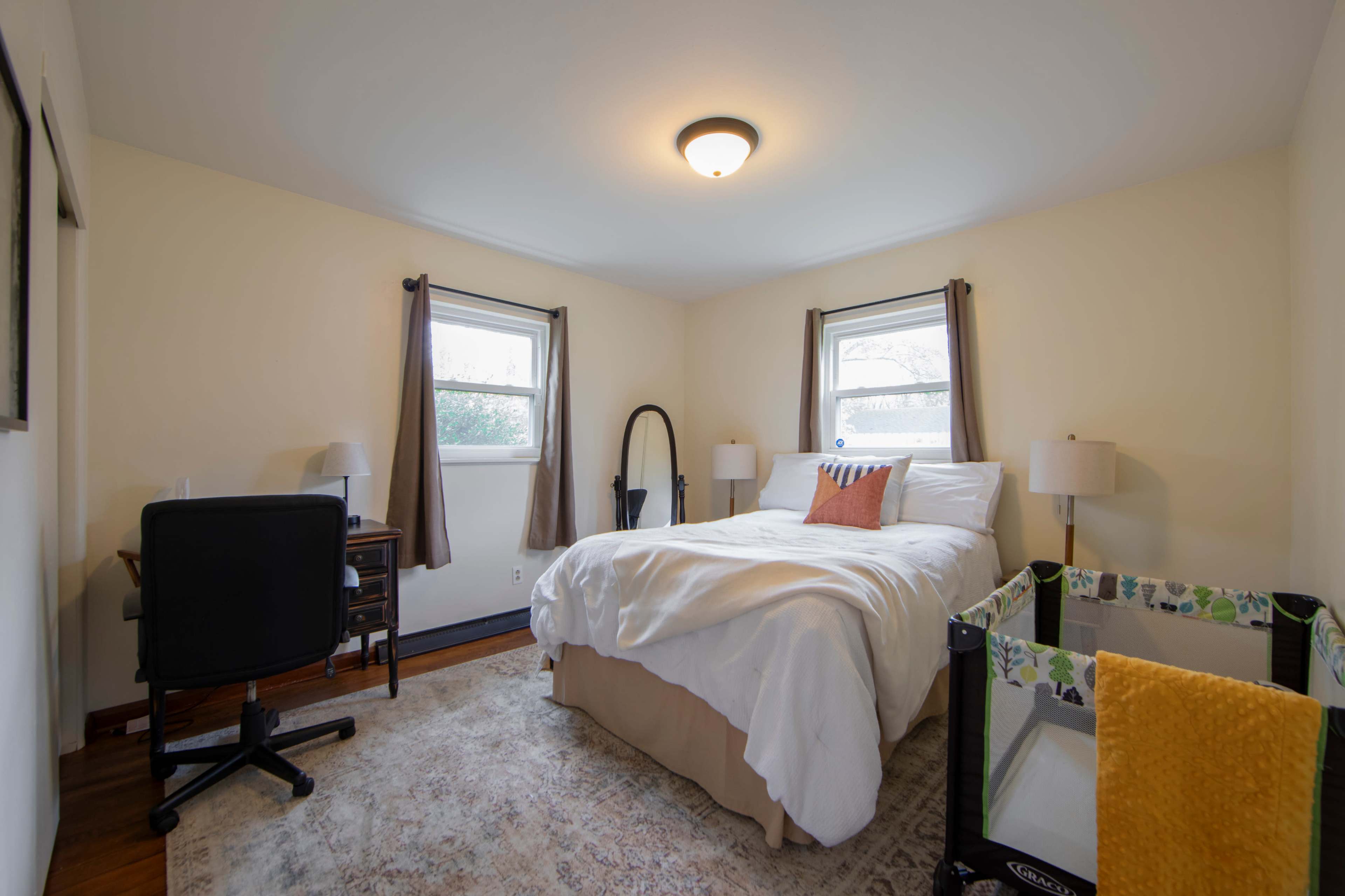 The image shows a bedroom with a bed, a desk and chair, a playpen, and two windows with curtains.
