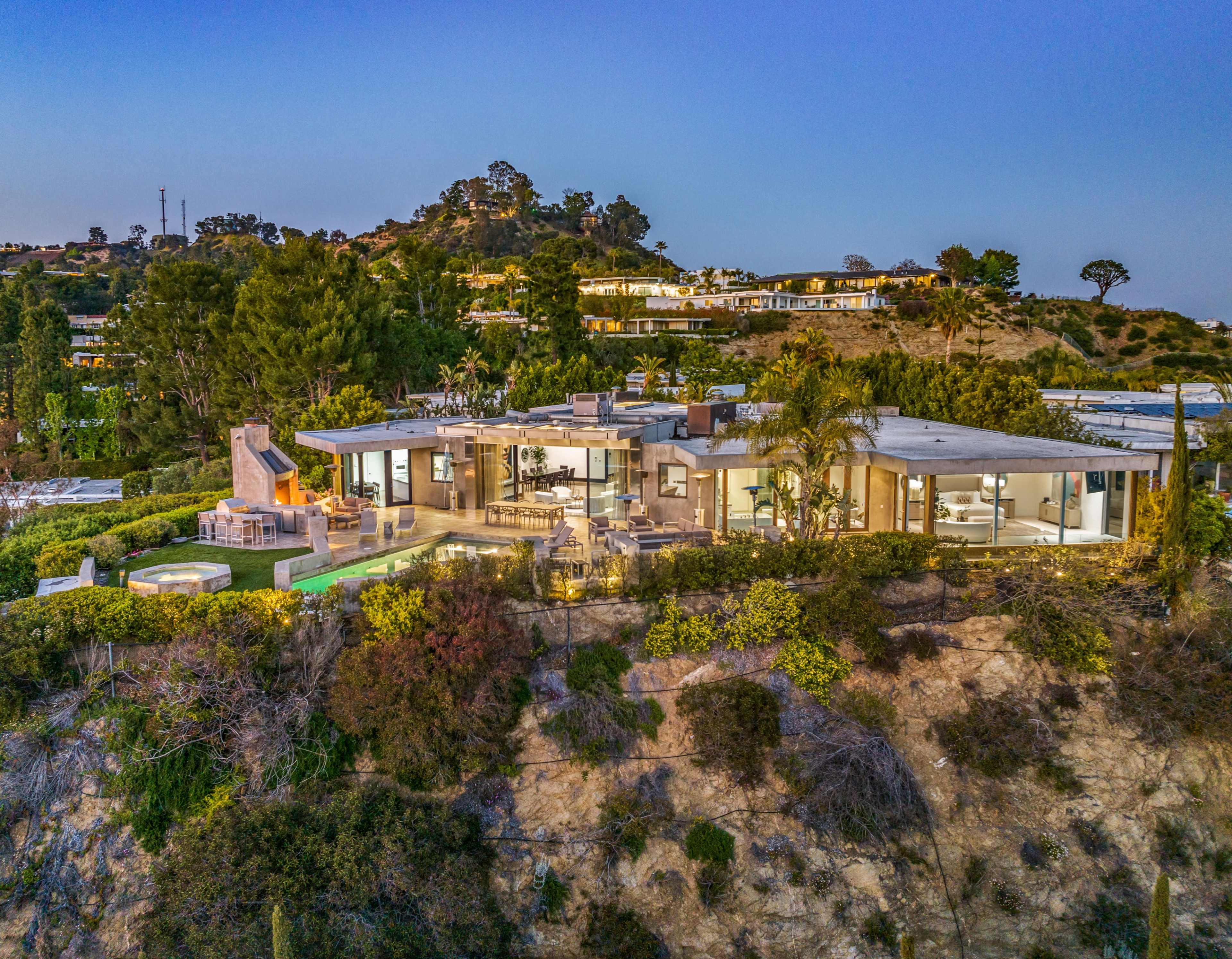 A modern house is situated on a hillside, featuring a spacious patio and large windows overlooking a lush landscape at sunset.