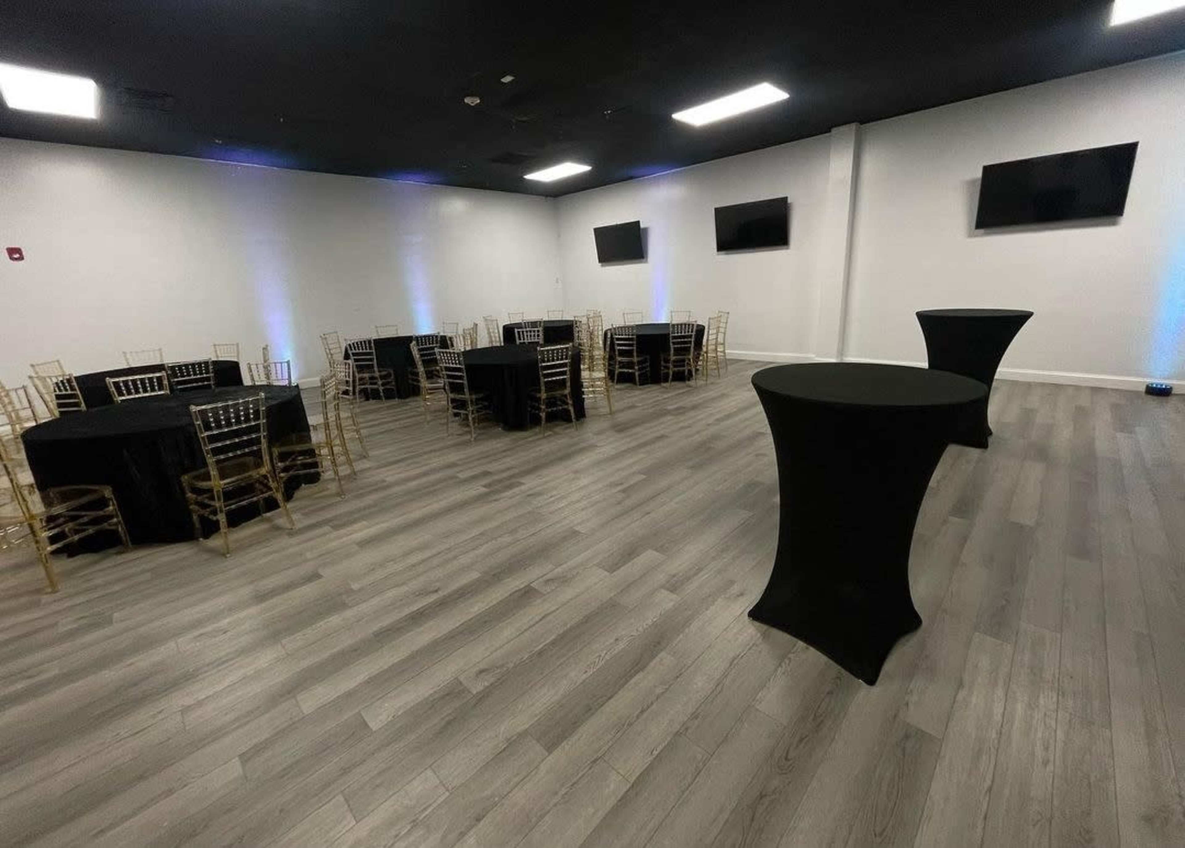 A spacious event room with several round tables covered in black cloth and high-top tables in the foreground, against a backdrop of white walls and black flooring.