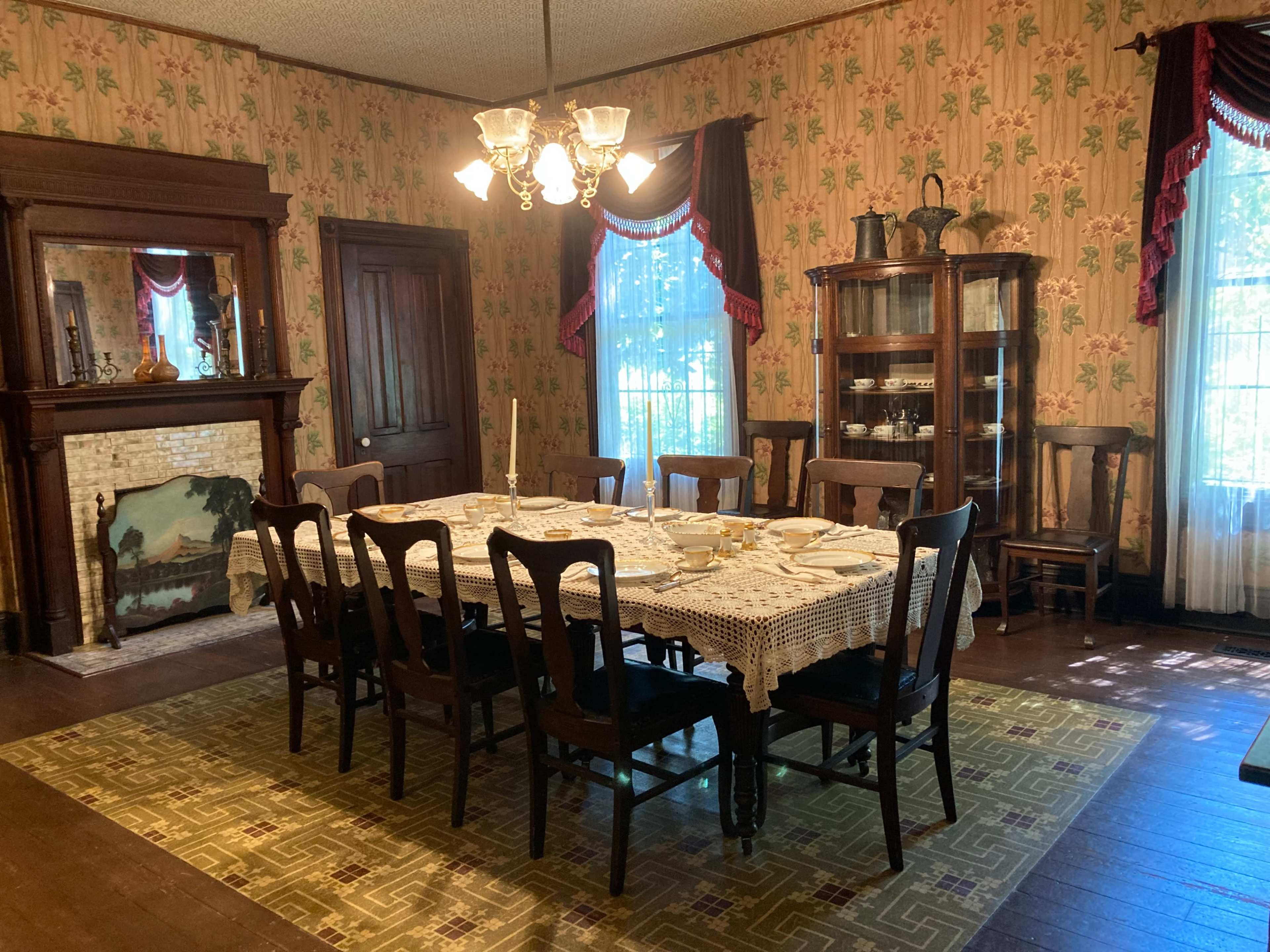 A formal dining room with a large table set for a meal, surrounded by wooden chairs, with a vintage fireplace and a china cabinet in the background.