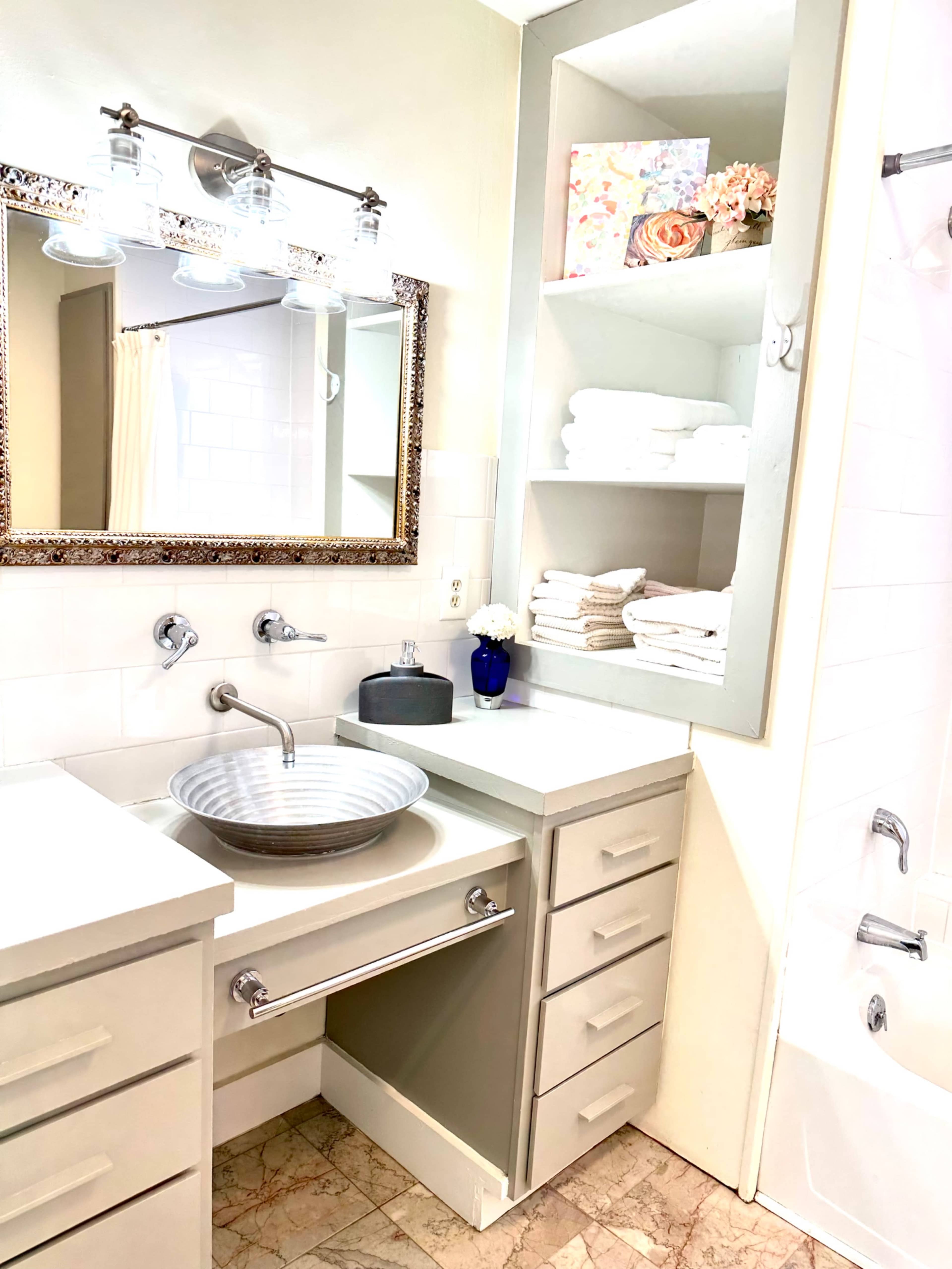 The bathroom features a modern sink with a striped bowl, surrounded by cabinets and shelves holding neatly stacked towels.