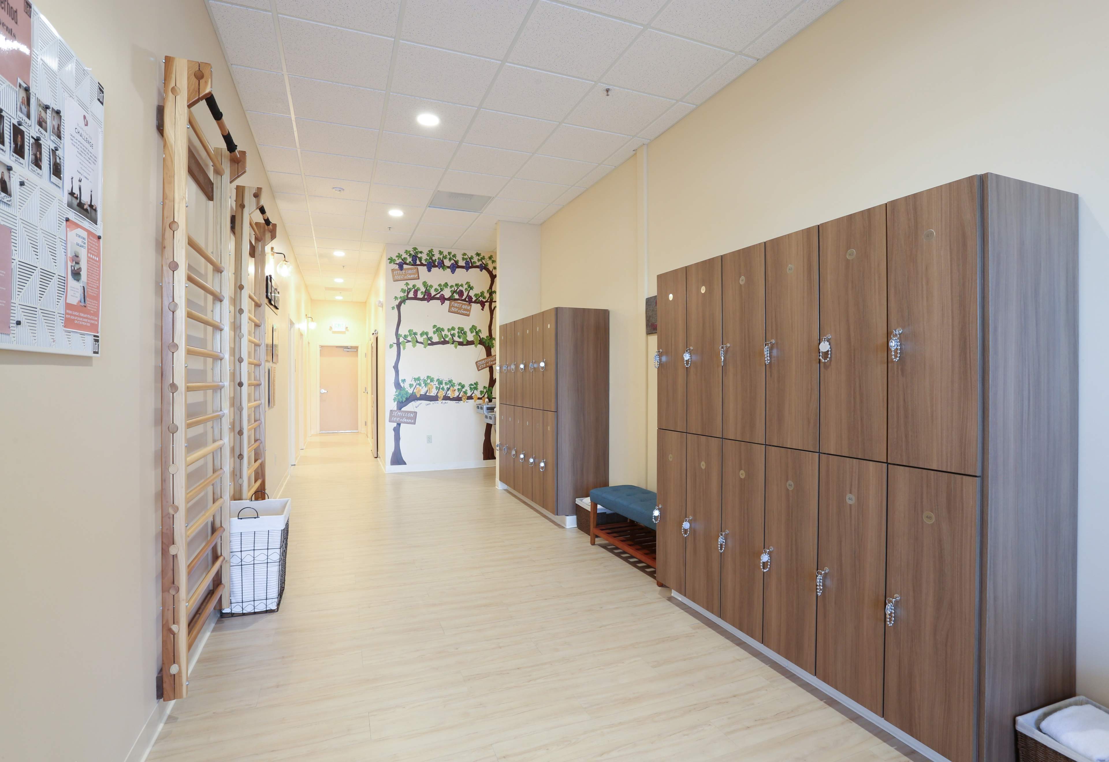 A corridor with wooden lockers on one side and a wall decorated with colorful artwork on the other, leading to a doorway at the far end.