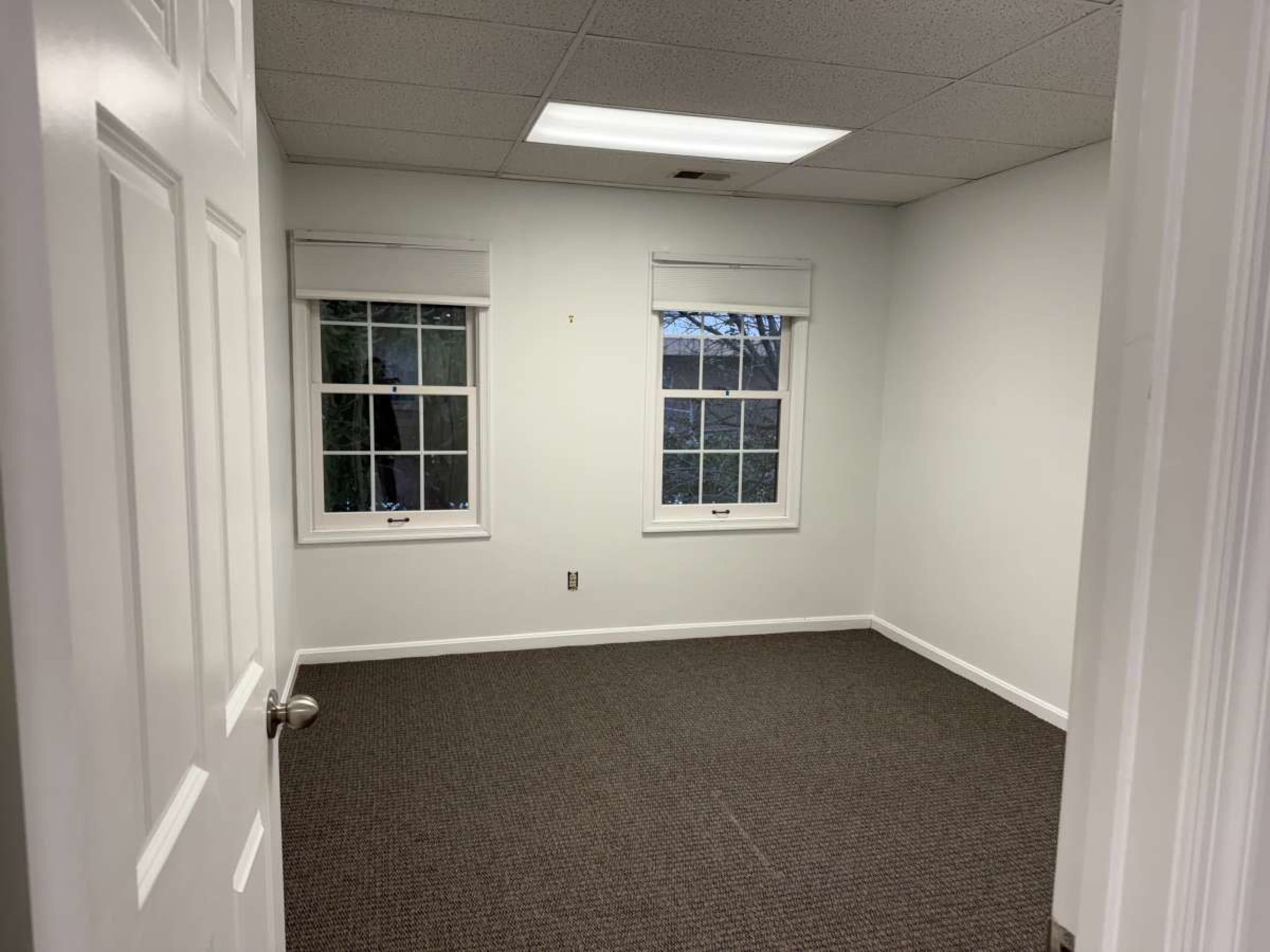 The image shows a vacant room with two windows and beige carpeted flooring.