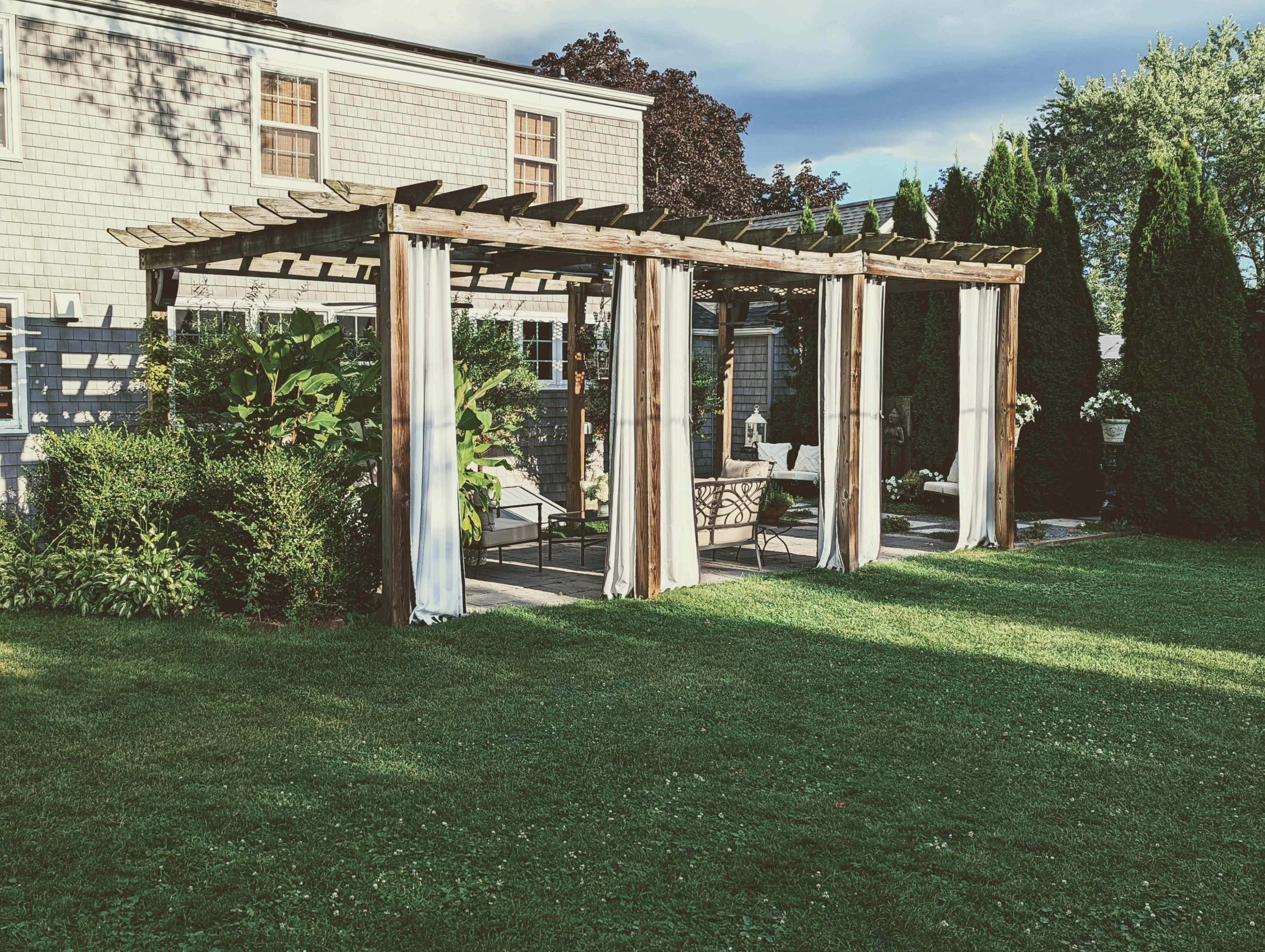 A wooden pergola with draped fabric curtains stands in a grassy backyard, providing a shaded seating area.