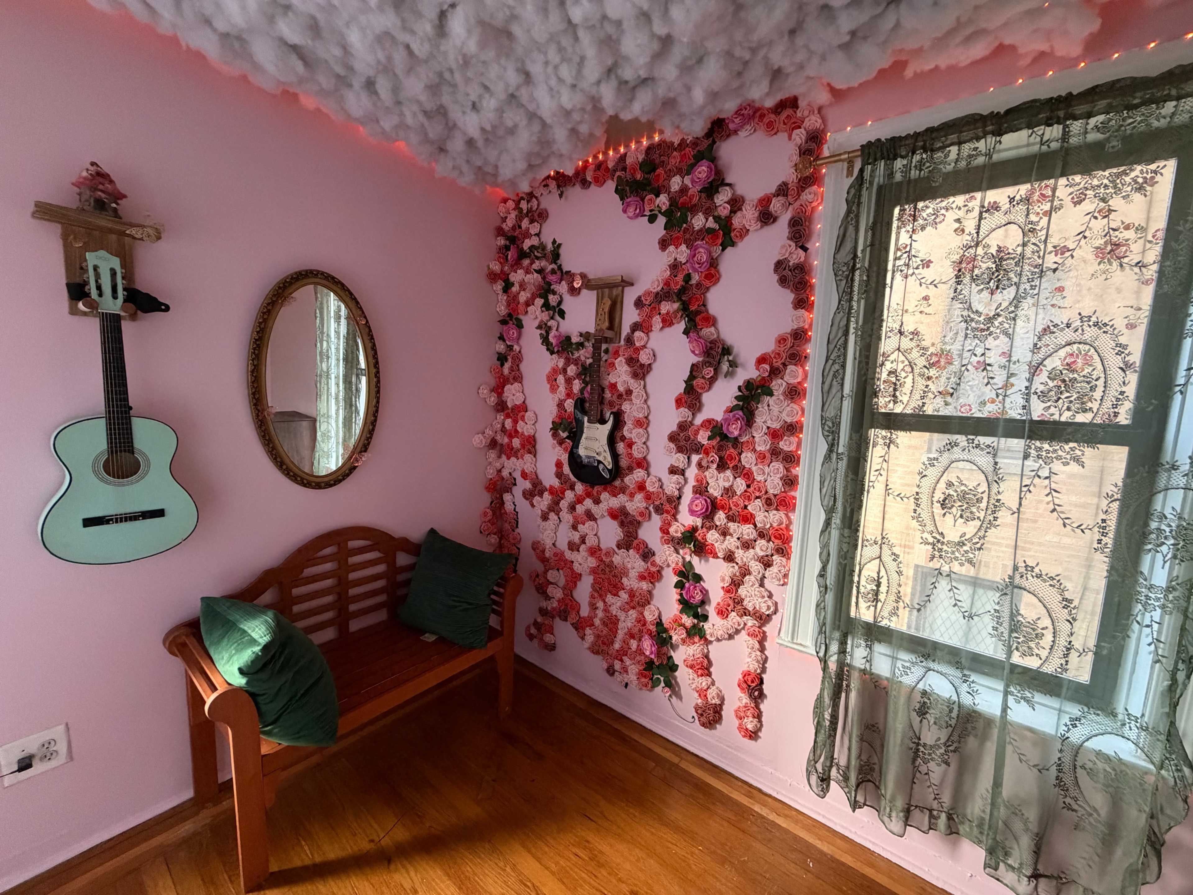 The room features a pink wall adorned with floral decorations, a mirror, a green acoustic guitar, and a wooden bench under a window with a patterned curtain.
