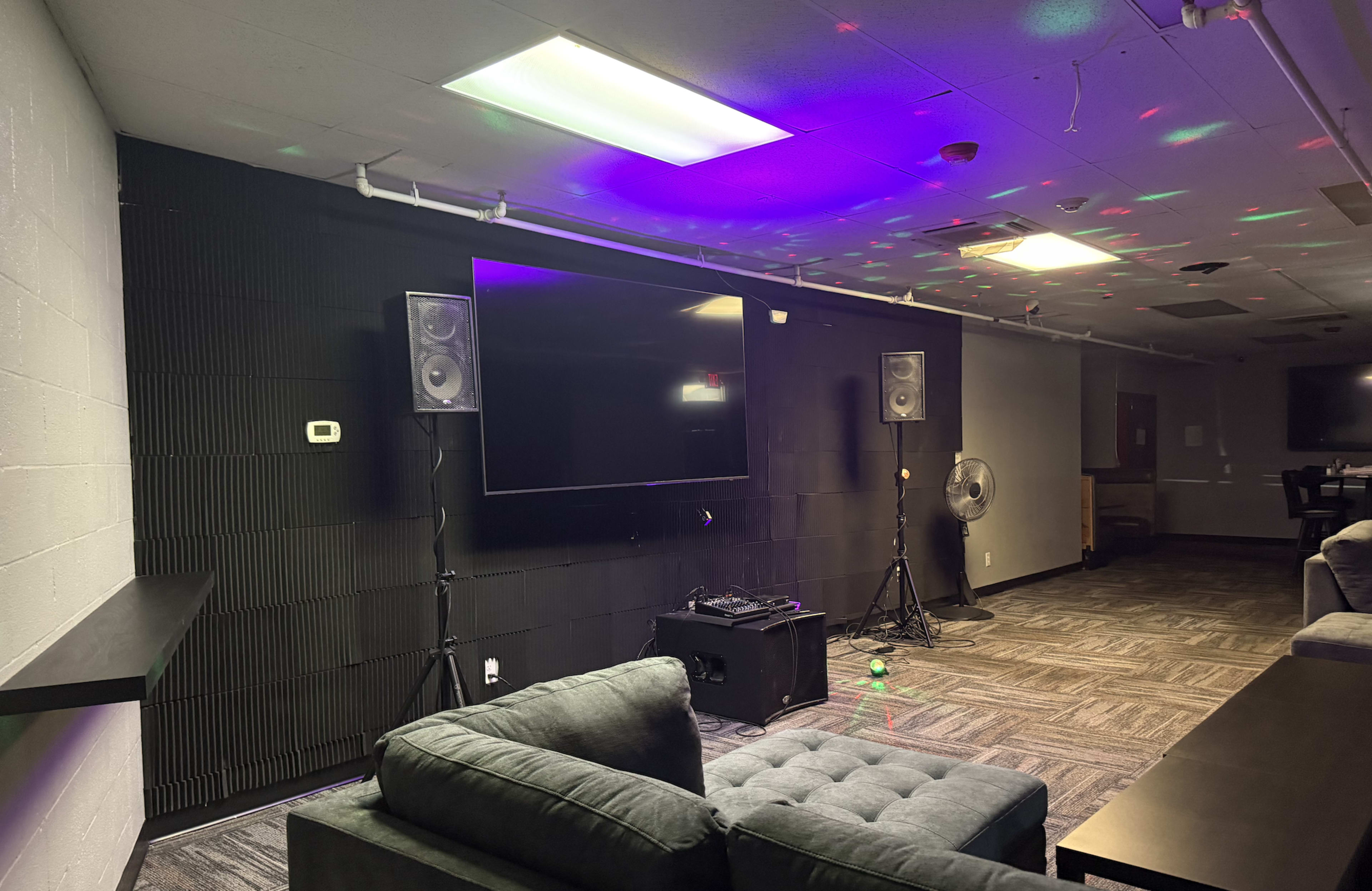 The image shows a lounge area with a large television mounted on a black wall, flanked by two speakers and a fan, with colorful lights reflecting on the ceiling.