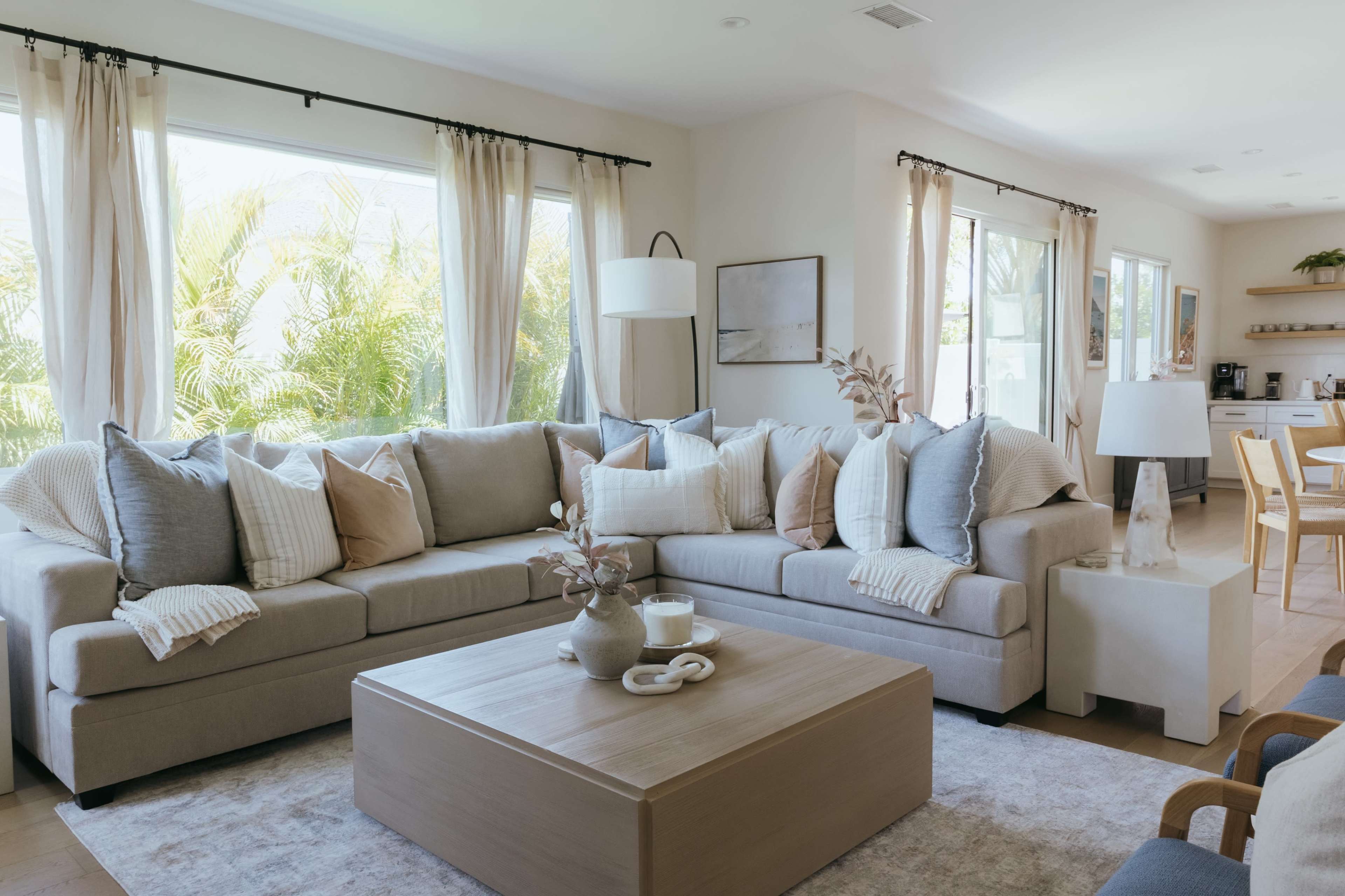 The image shows a cozy living room with a large sectional sofa adorned with various cushions, a wooden coffee table in the center, and large windows letting in natural light.