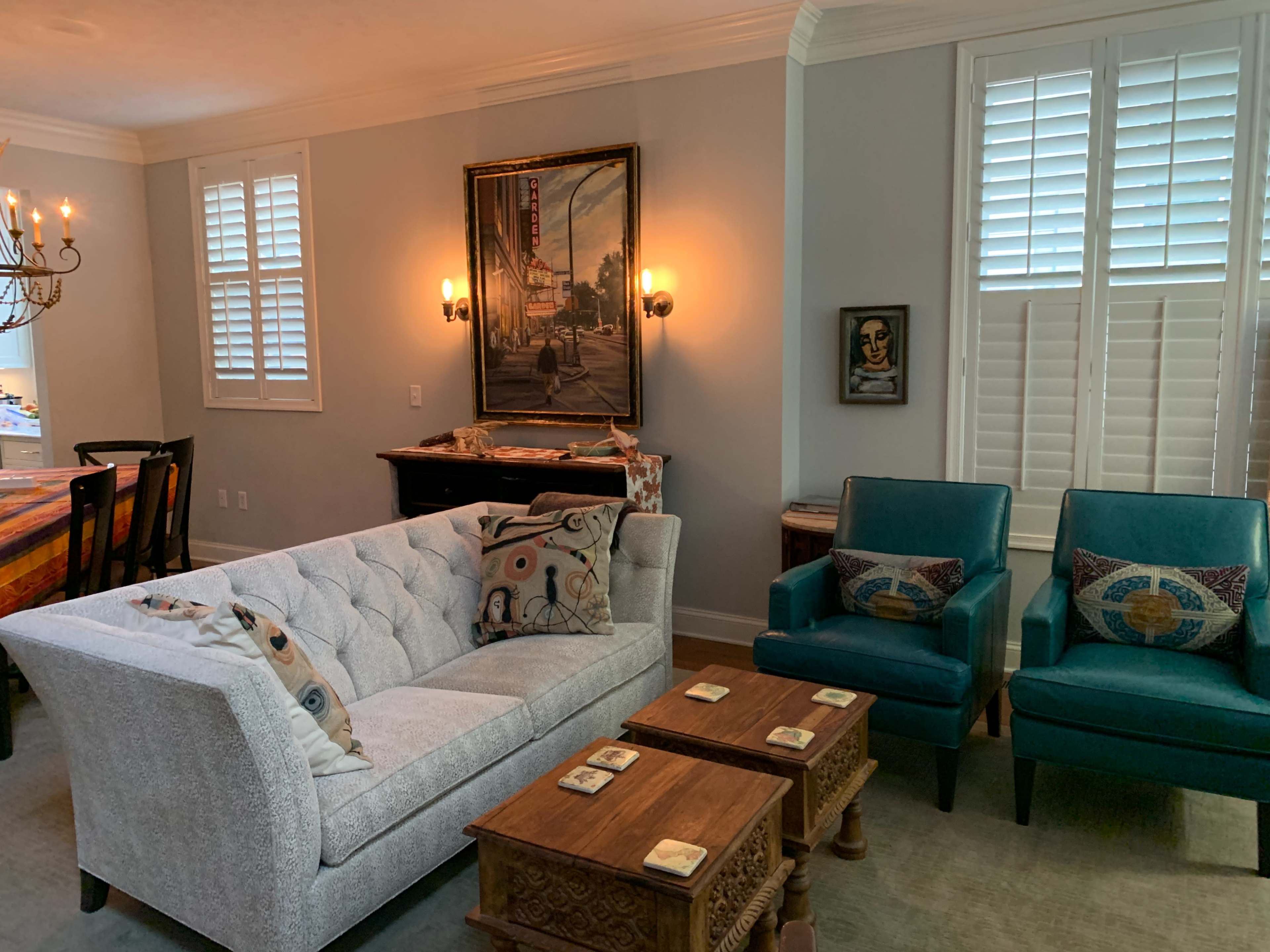 A living room with a gray tufted sofa, two teal armchairs, wooden coffee tables, and artwork on the walls.
