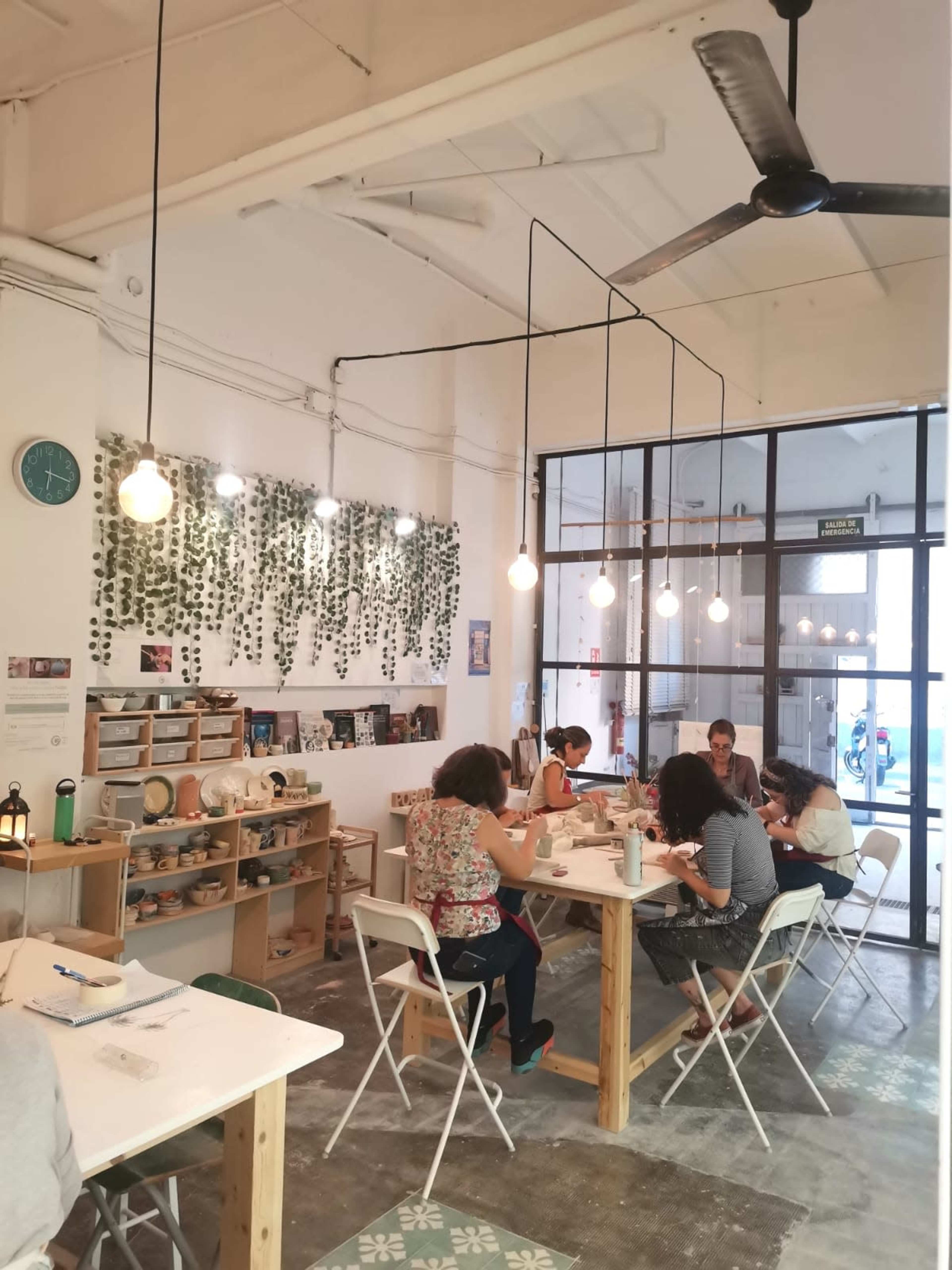 The image shows a bright, open studio space with people sitting at tables, engaged in a craft activity, under pendant lights and surrounded by greenery on the walls.