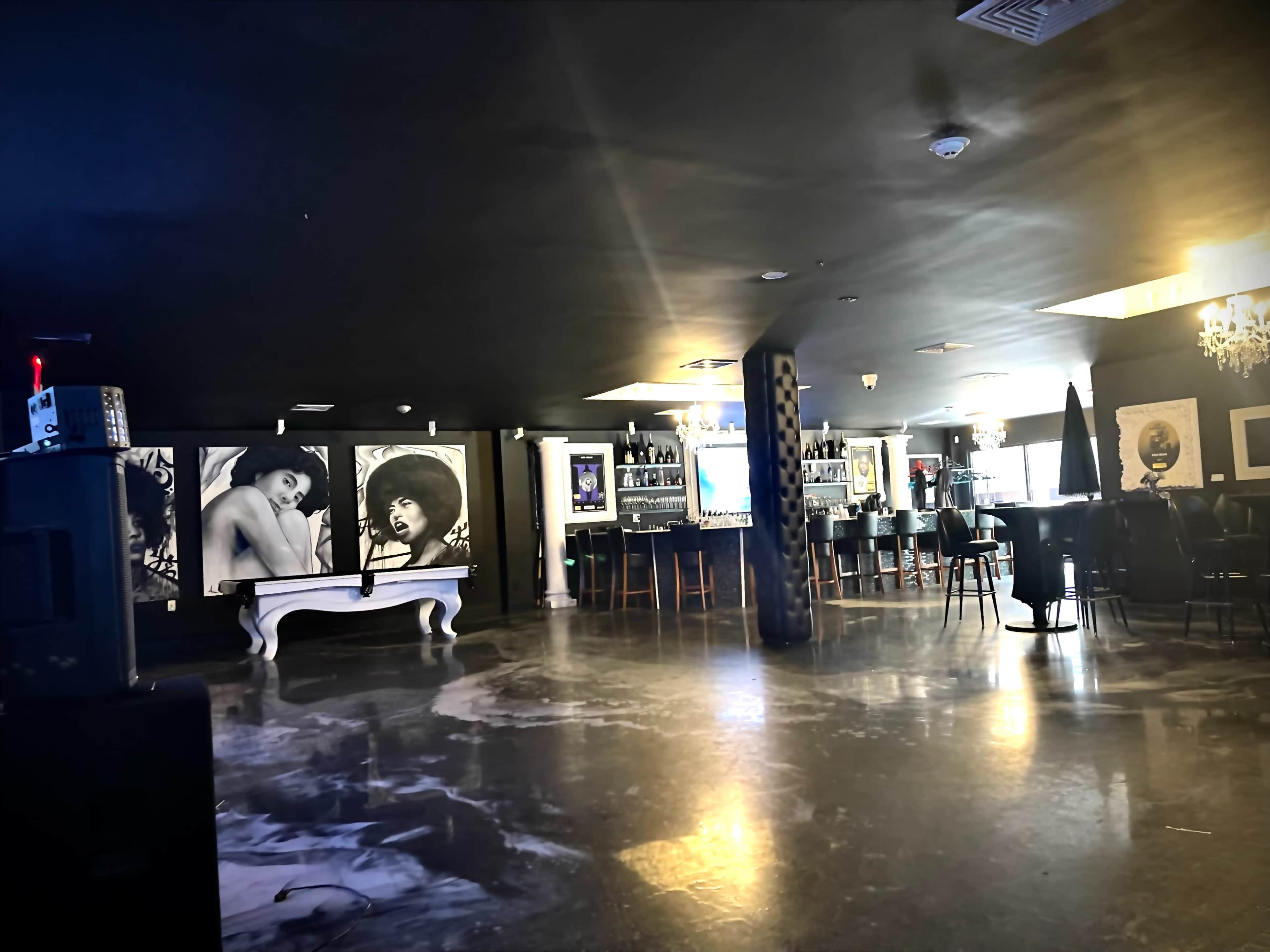 The interior of a dimly lit bar features large black-and-white portraits on the walls and a long counter with stools.